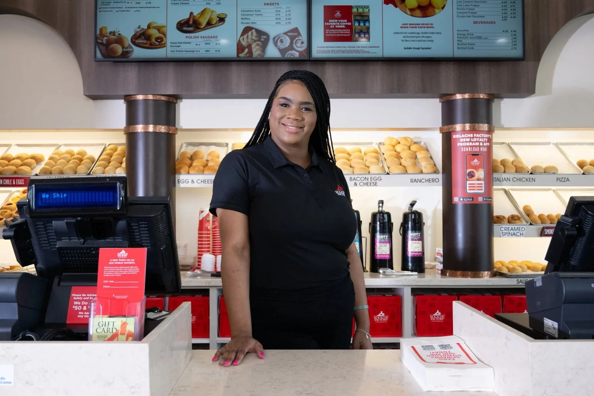 A smiling woman in a black uniform standing behind the counter of a Kolache Factory.