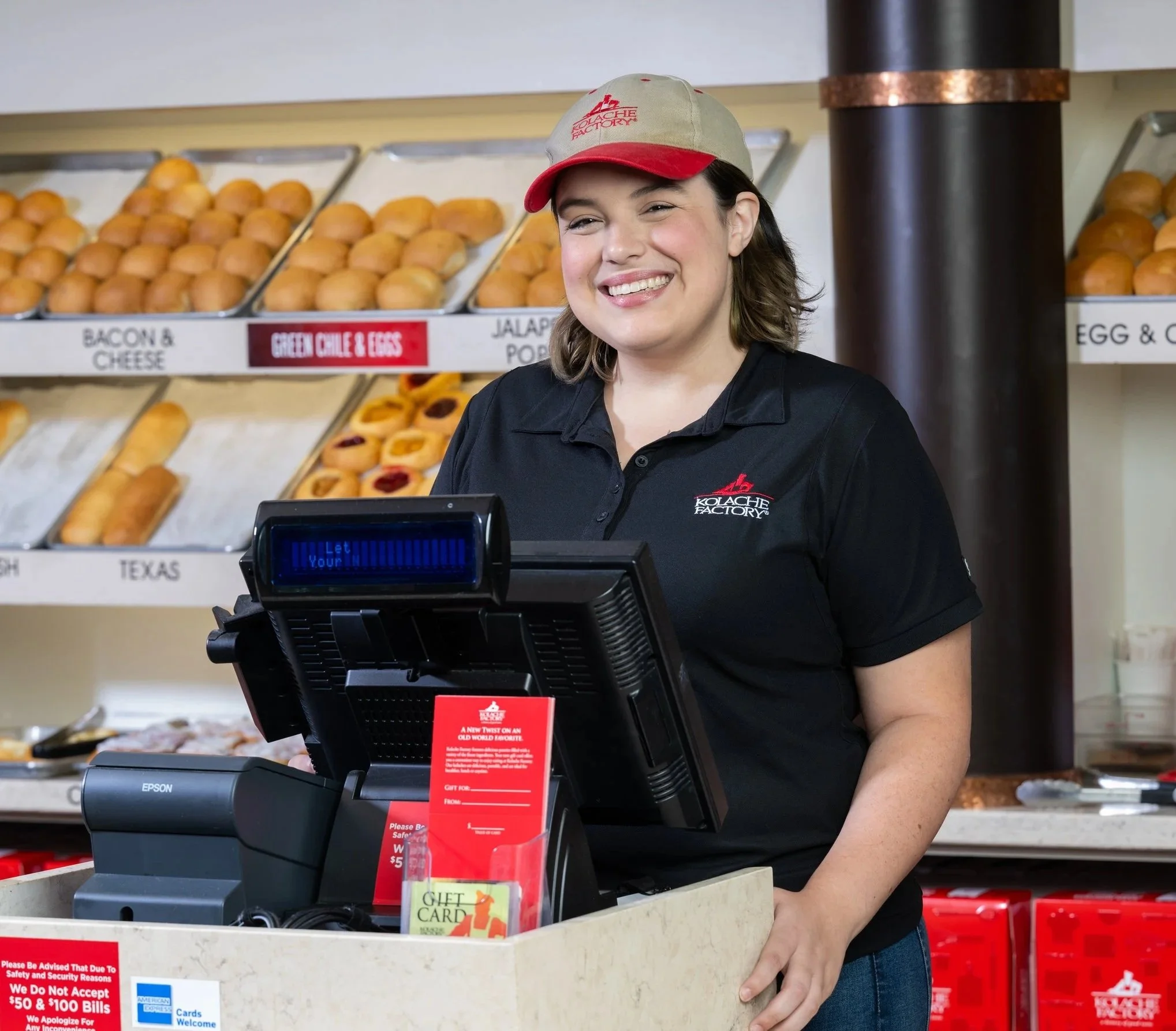A woman in a black polo shirt and a beige and red cap smiling at a Kolache Factory bakery counter.