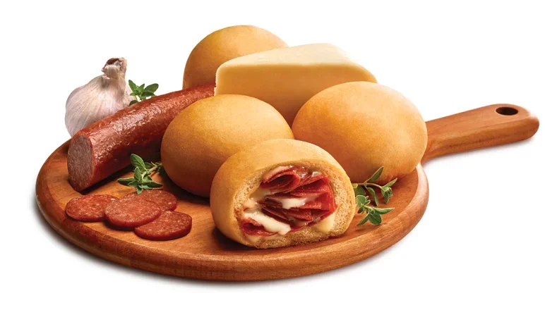 A wooden serving board with three Kolaches filled with pepperoni, cheese, and tomato sauce, accompanied by slices of salami, a garlic bulb, and some herbs.