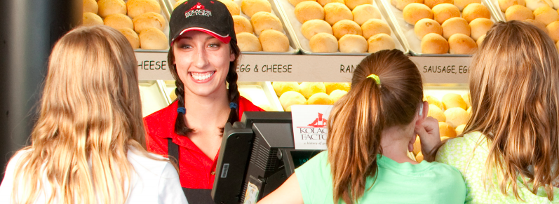 A smiling female employee at aKolache Factory cafe counter surrounded by children, with shelves of Kolaches behind her.