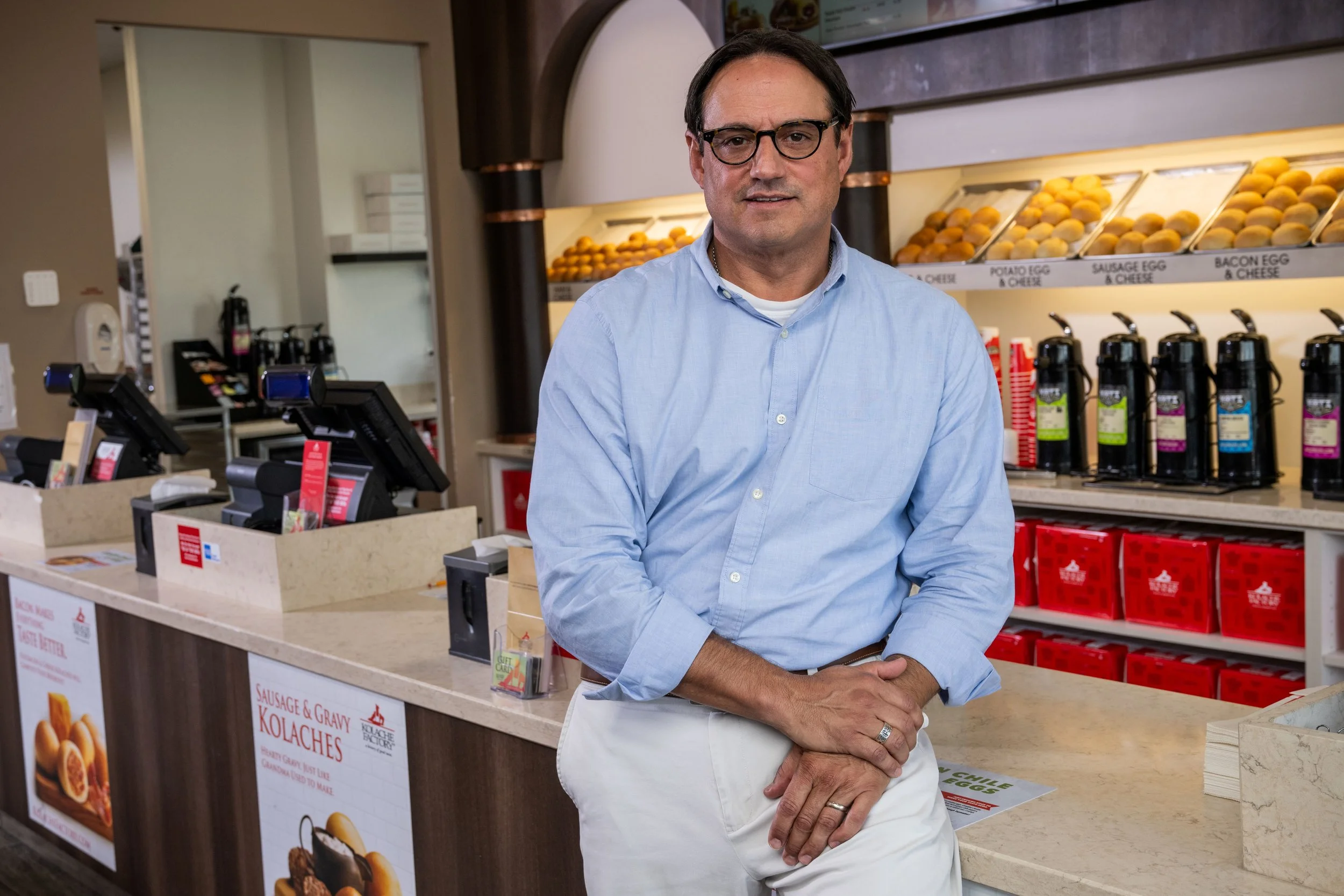 A man with glasses wearing a light blue button-down shirt standing in front of a Kolache Factory cafe counter.