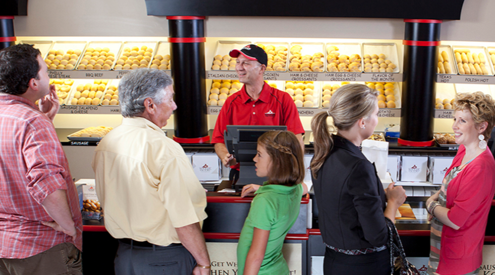 People standing in line at a Kolache Factory cafe counter, with various Kolaches on display behind the cashier.