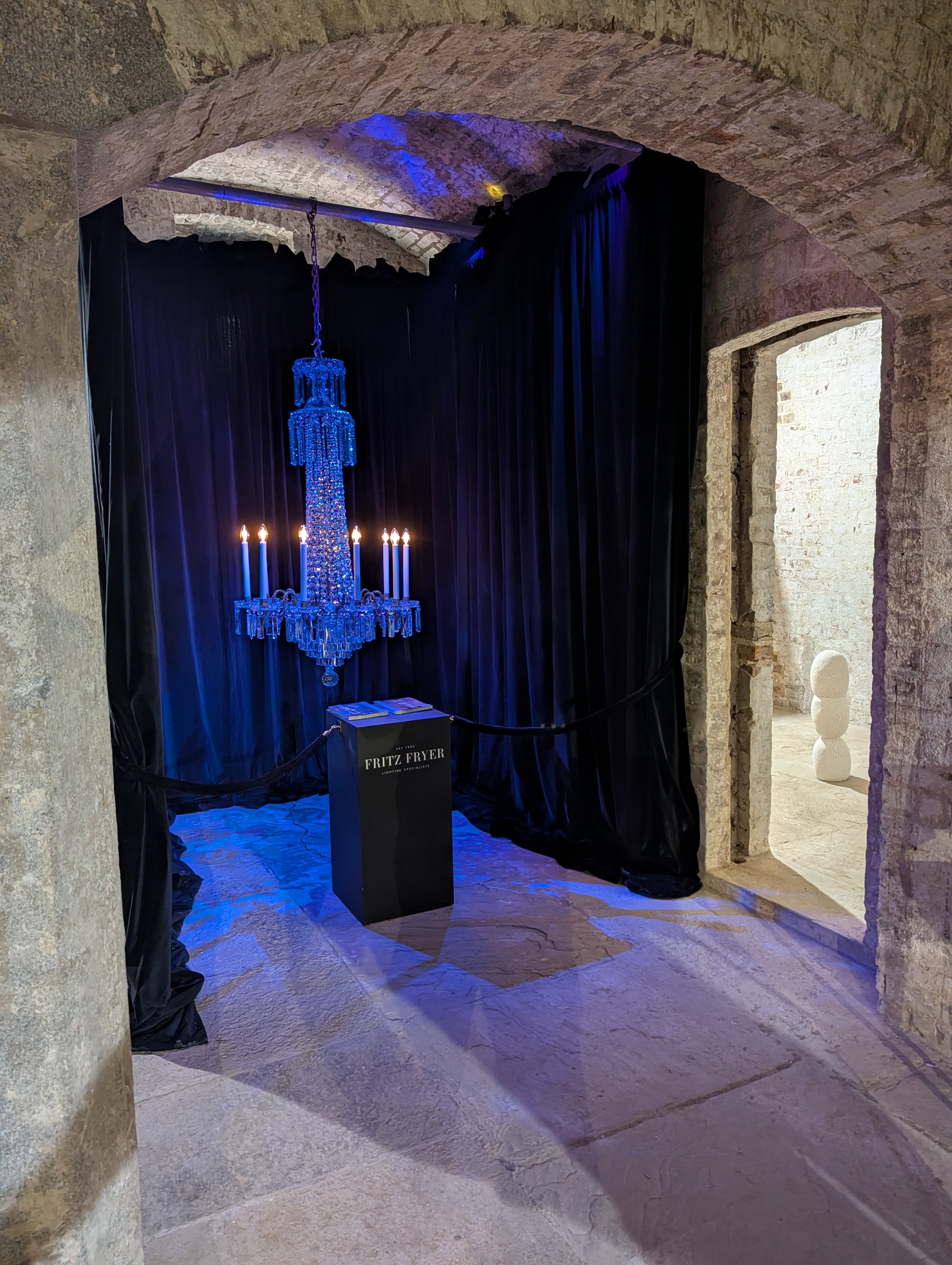 Elaborate blue chandelier hanging in front of black curtains in a stone-walled room, with a sign that says 'Fritz Fryer Lighting Specialists'.