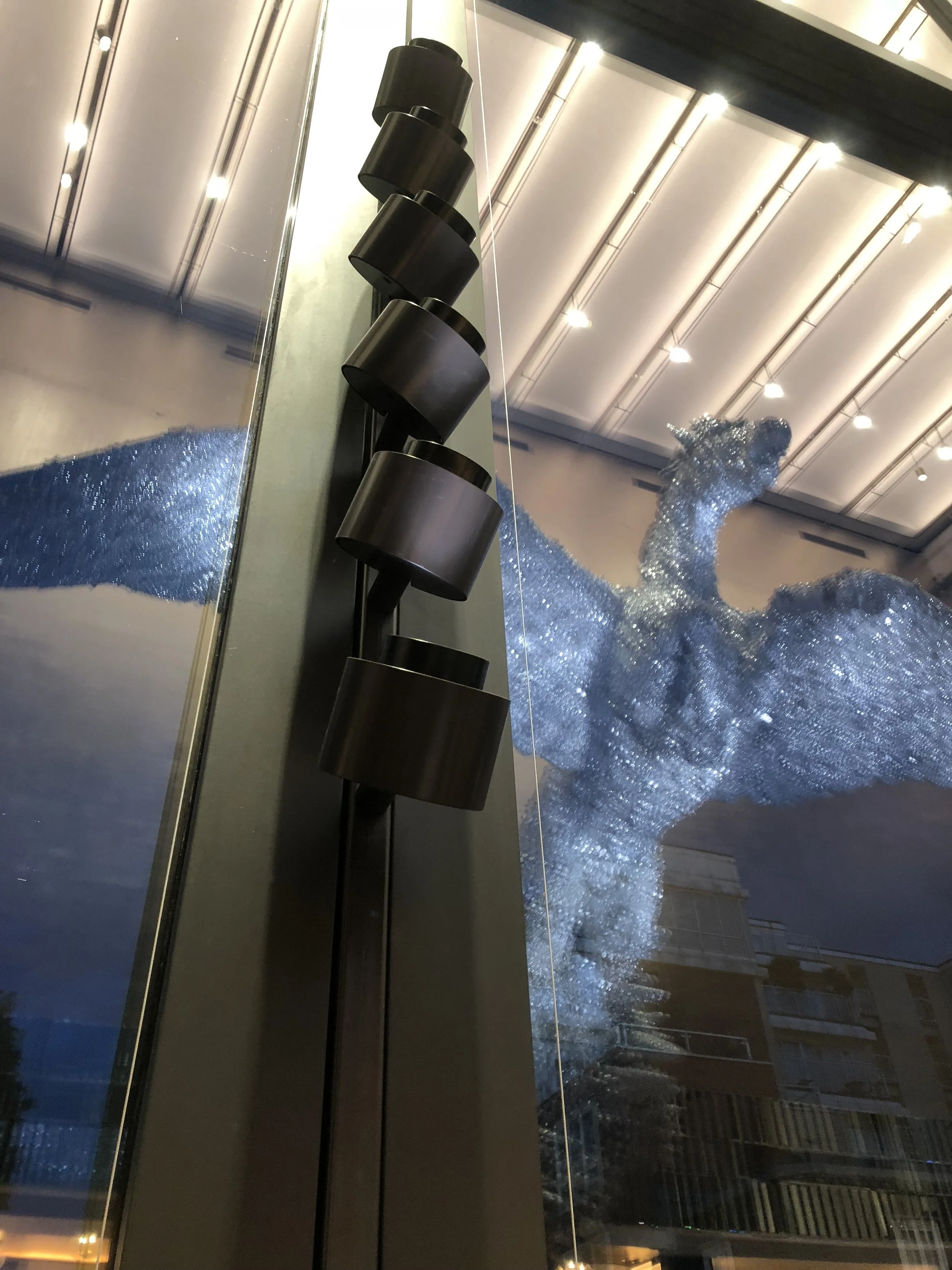 Reflection of a large, glittering sculpture resembling an animal, possibly a dog, in a glass window inside a modern building with a high ceiling and hanging lights. The sculpture appears to be covered in shiny, reflective material.