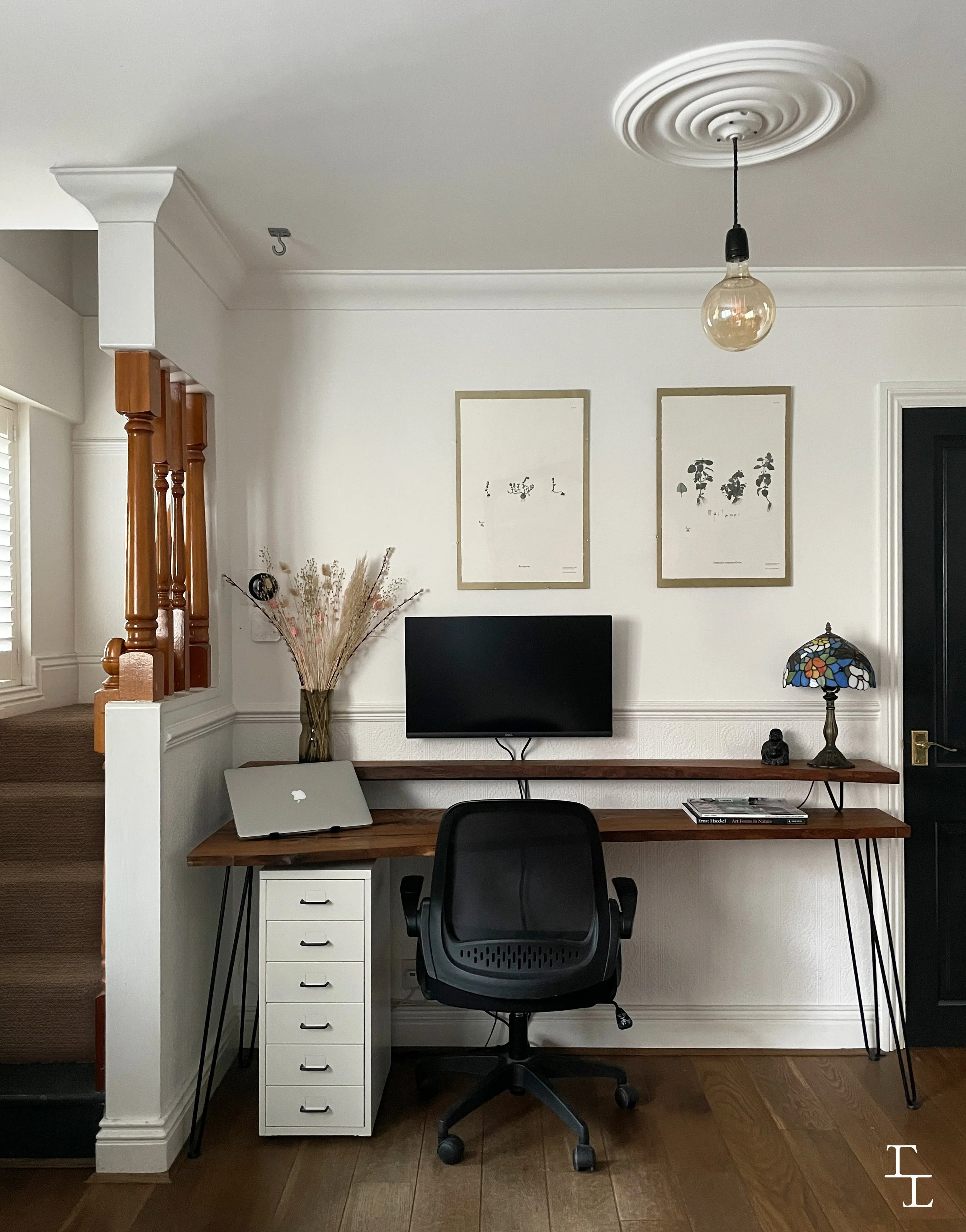 Home office setup with a wooden desk holding a MacBook laptop, a black office chair, a white mini drawer unit, a glass vase with dried flowers, a stained glass table lamp, a flat-screen monitor mounted on the wall, framed botanical prints, and a black Buddha statue.
