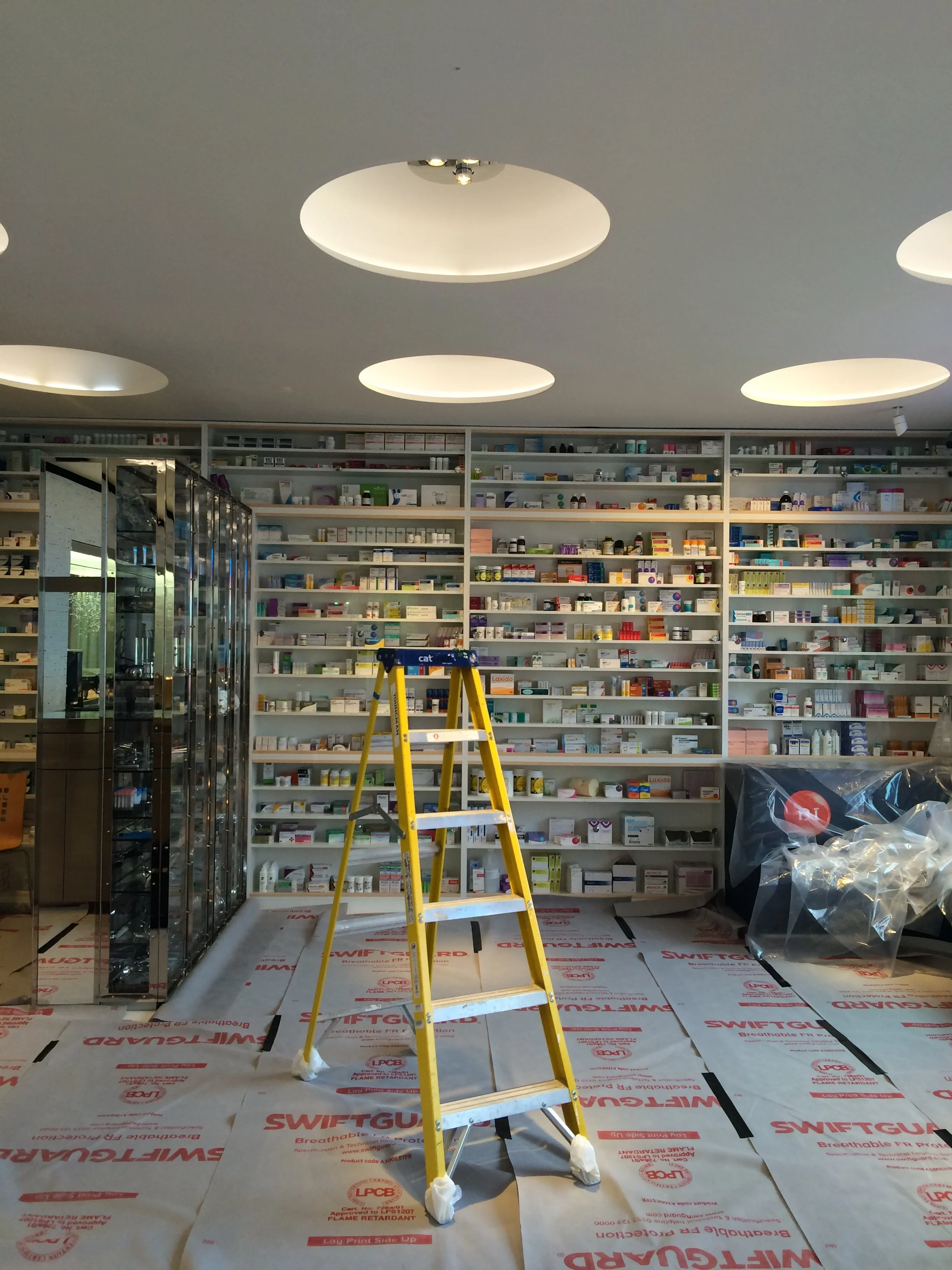 A pharmacy interior with shelves full of medication bottles and boxes, a yellow step ladder in the center, and a glass door to the left. The ceiling has round, recessed lights.