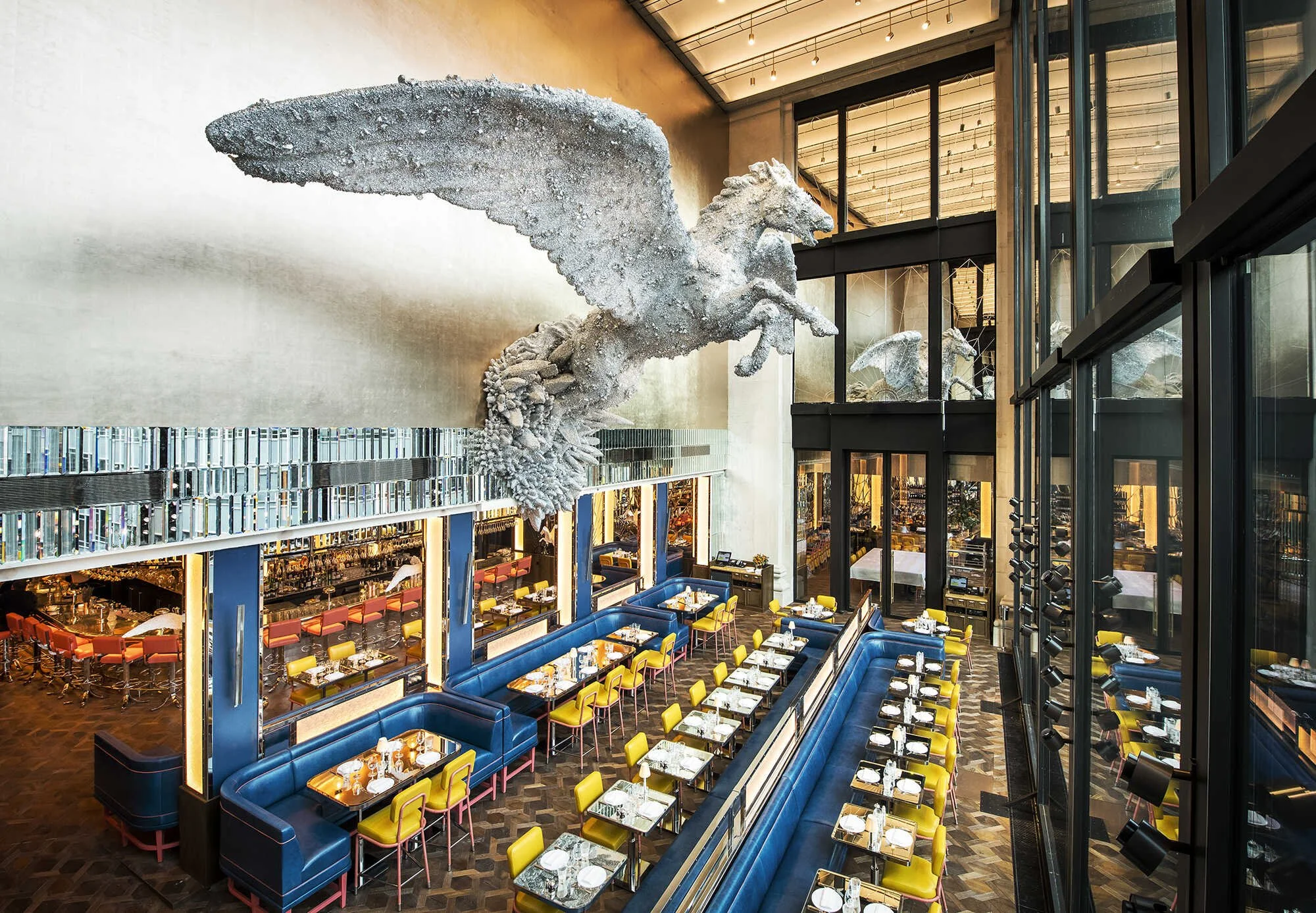 Multiple dining tables with yellow chairs and blue booths in a modern restaurant with floor-to-ceiling windows and an artistic sculpture of a Pegasus mounted on the wall.