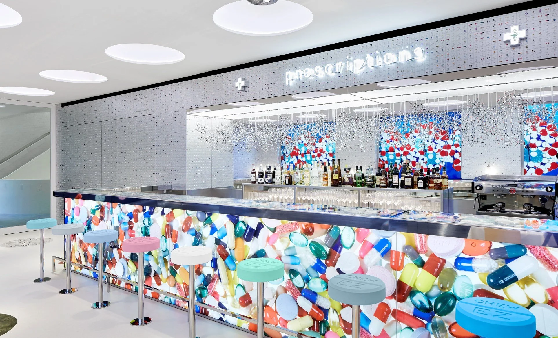 Modern pharmacy or bar area with a wall and bar front decorated with images of colorful pills and capsules. Shelves behind the bar display various bottles of alcohol. Artic-inspired lighting and colorful seating.