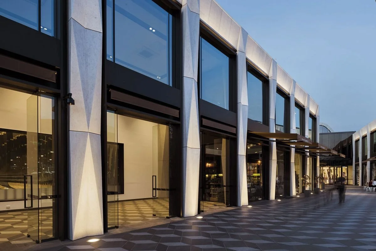 Modern shopping center or mall exterior with large glass windows and patterned pavement, during evening hours.