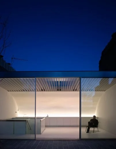Interior view of a modern, minimalist room with large glass windows, a person sitting alone, and a dark outdoor sky.