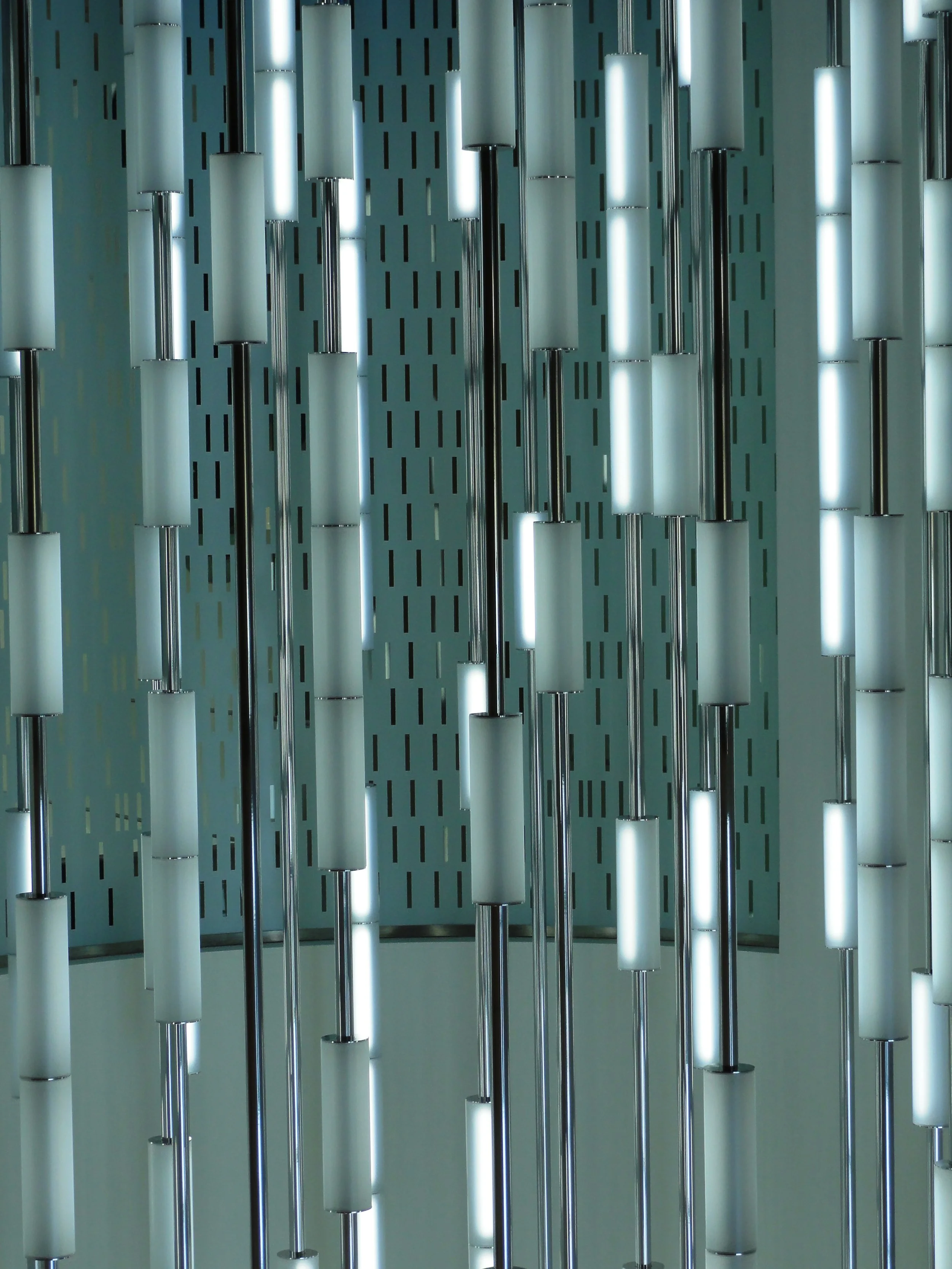 Vertical illuminated LED light fixtures on black rods, arranged in parallel rows on a wall with a perforated pattern background.