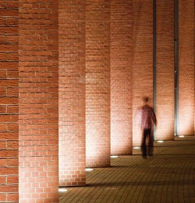 Person walking along a brick wall illuminated by ground lights at night.