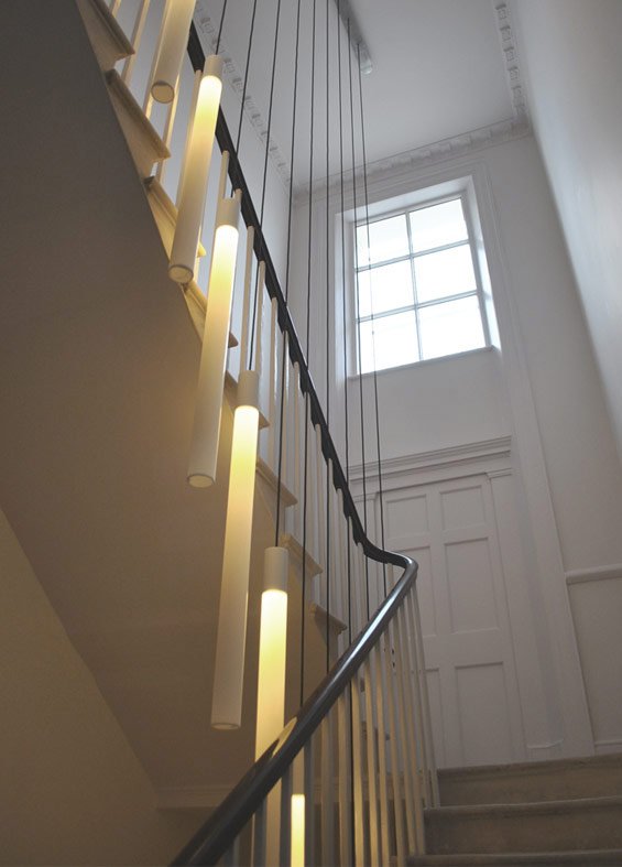 A staircase with modern cylindrical wall-mounted lights along the side, black handrail, white walls, and a large window at the top letting in natural light.