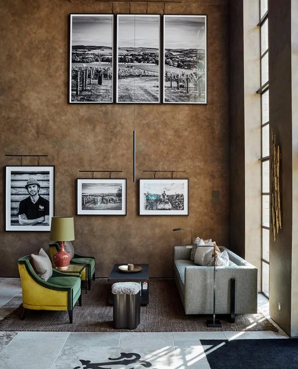 Living room with a textured brown wall, large windows on the right, and framed black-and-white photographs of landscapes and a person on the wall. There is a green and yellow armchair, a grey sofa, a small black table, a lamp, and decorative pillows.