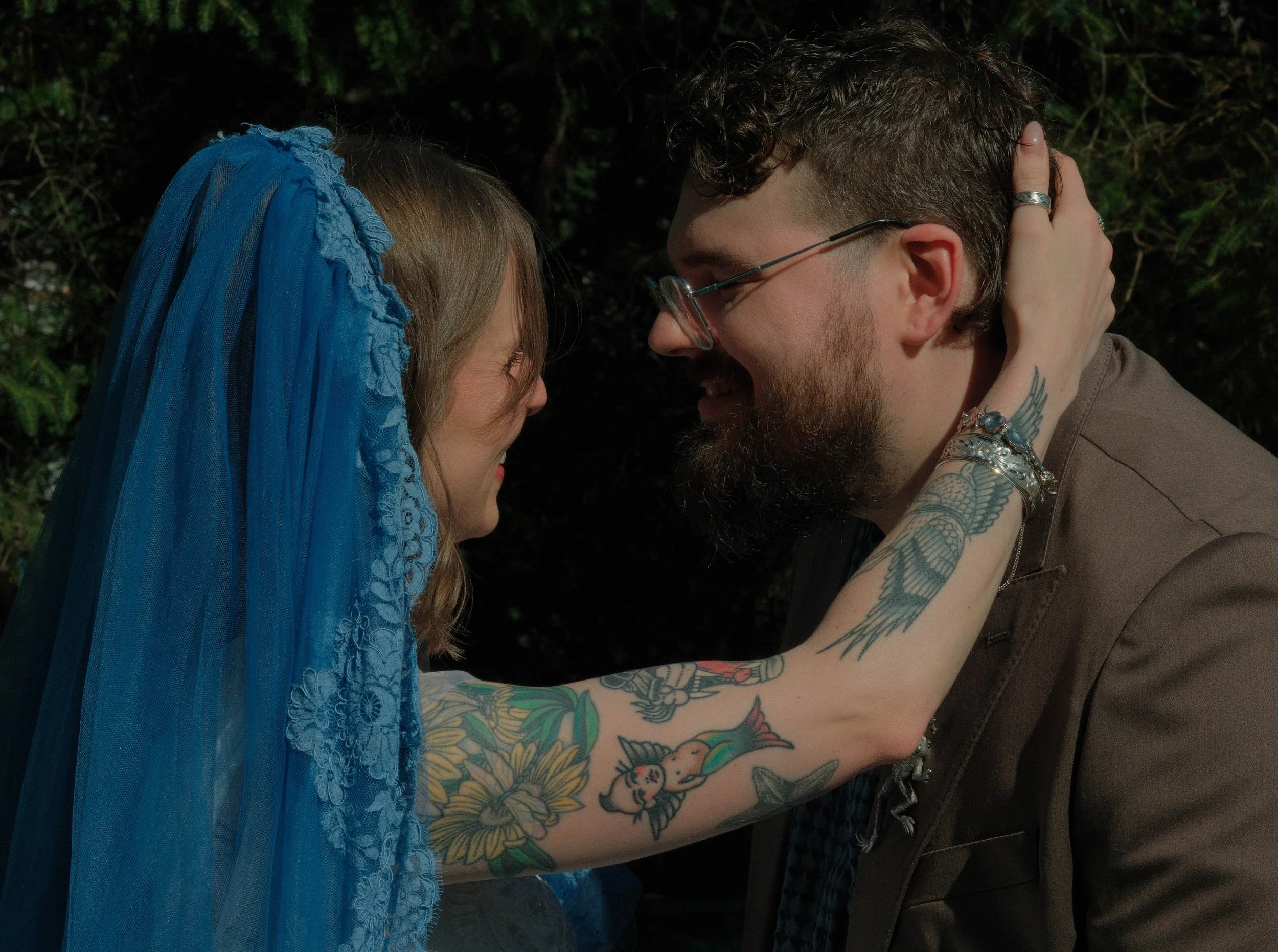 A close-up of a couple touching foreheads, holding each other's faces, both smiling. The woman wears a blue headscarf, and the man has tattoos on his arm.