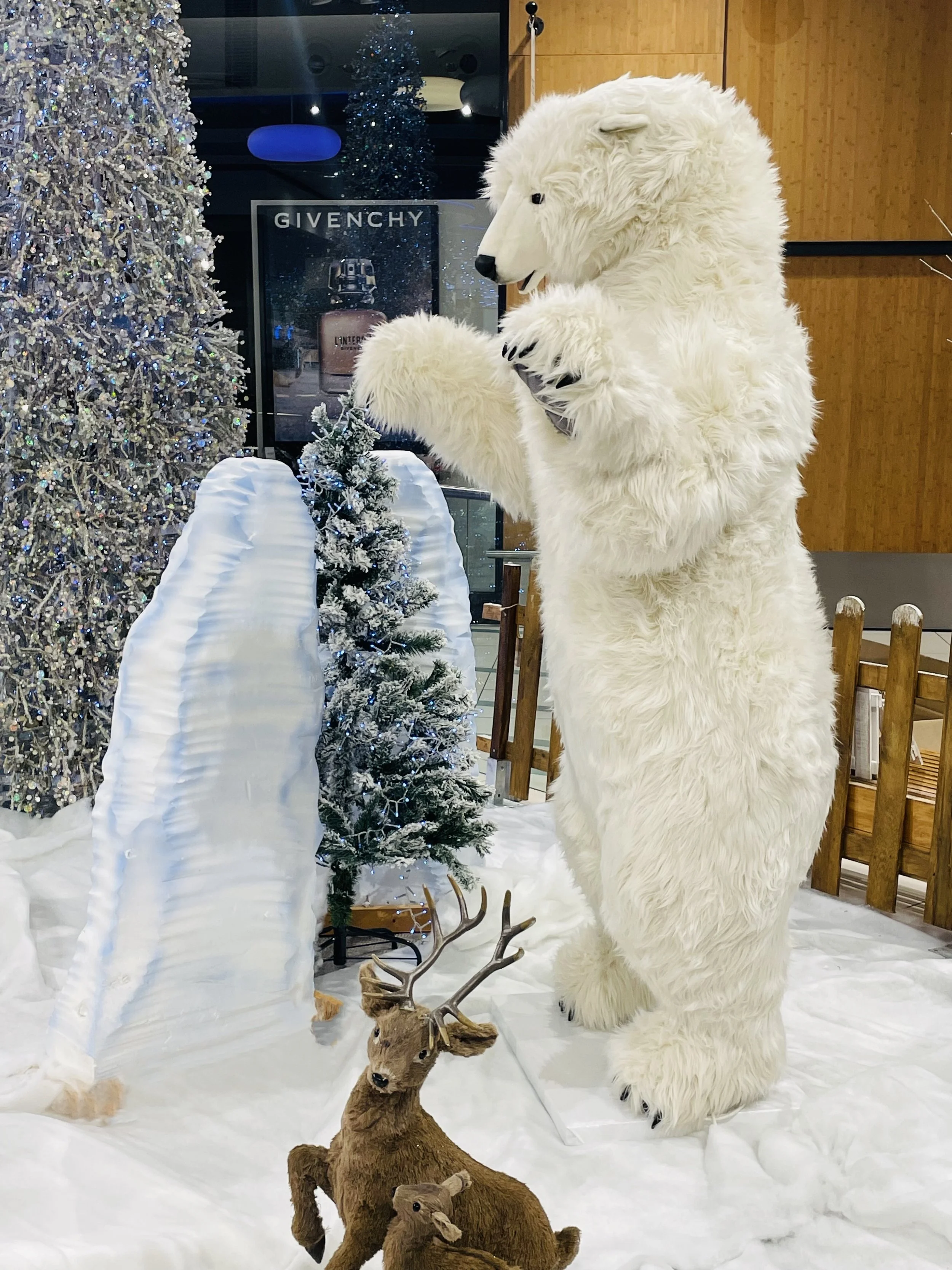 Décoration intérieure de Noël comprenant des sculptures d'ours polaires blancs automates, un sapin décoré, des bonshommes de neige, des rennes et des branches blanches, le tout entouré d'une petite clôture en bois. dans un centre commercial.