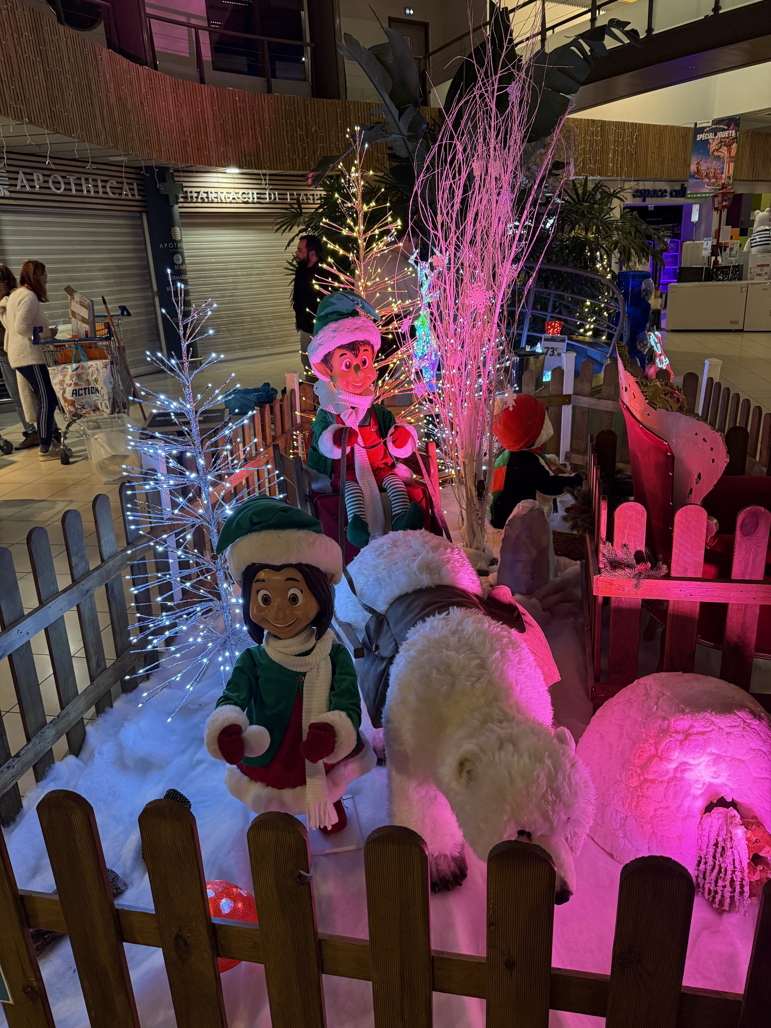 Décoration de Noël intérieure mettant en scène une grande figurine d'elfe vêtue d'un costume rouge et d'un bonnet de Père Noël, entourée d'arbres illuminés, d'éclairages roses et violets, d'un enclos avec des ours polaires automates en peluches.