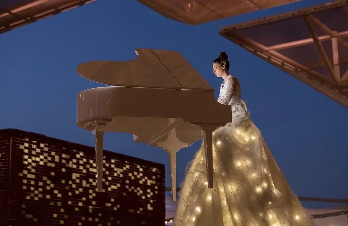Une femme en robe longue illuminée joue du piano blanc sur un toit lors de la nuit