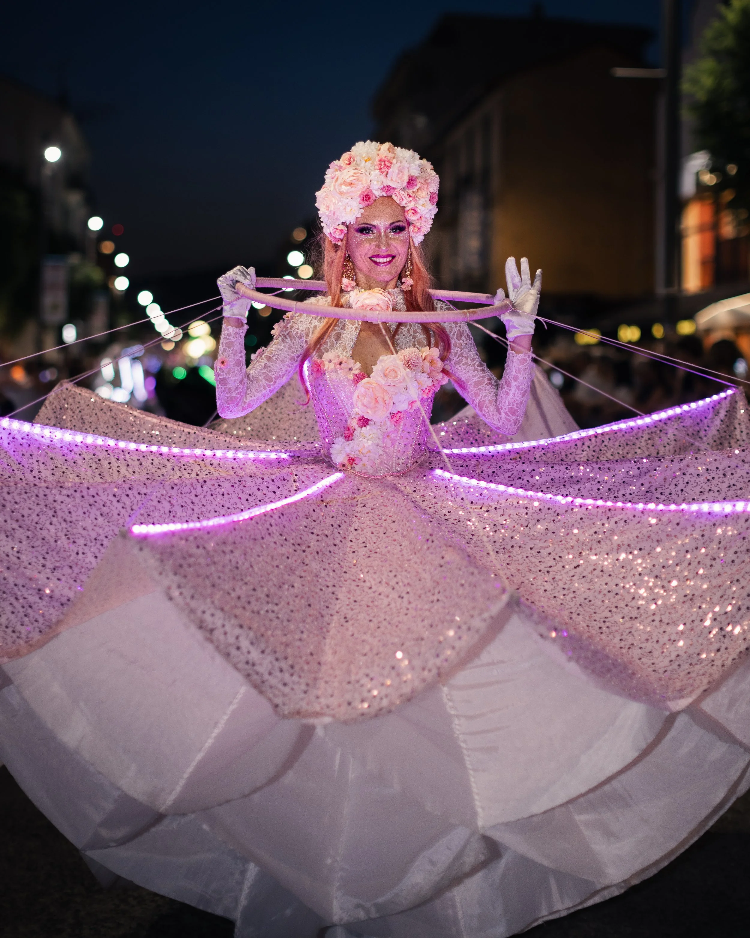 Personne vêtue de rose, coiffée d'une grande couronne de fleurs et portant une robe à paillettes, souriant la nuit lors d'un défilé ou d'une fête, entourée de lumières.