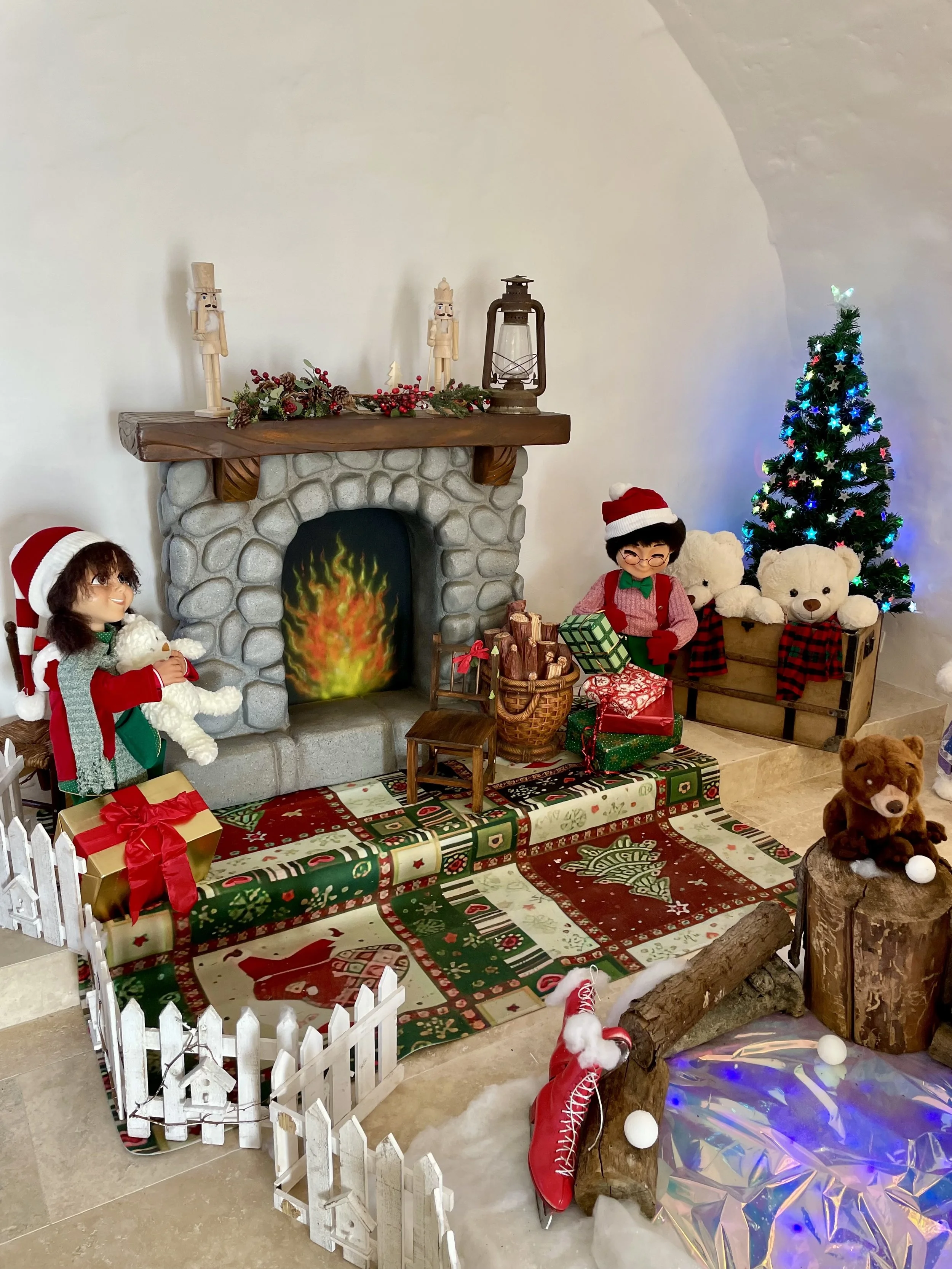 Scène de Noël chaleureuse avec un sapin décoré, des peluches, des poupées elfes, une cheminée avec un feu, des cadeaux et des décorations festives.