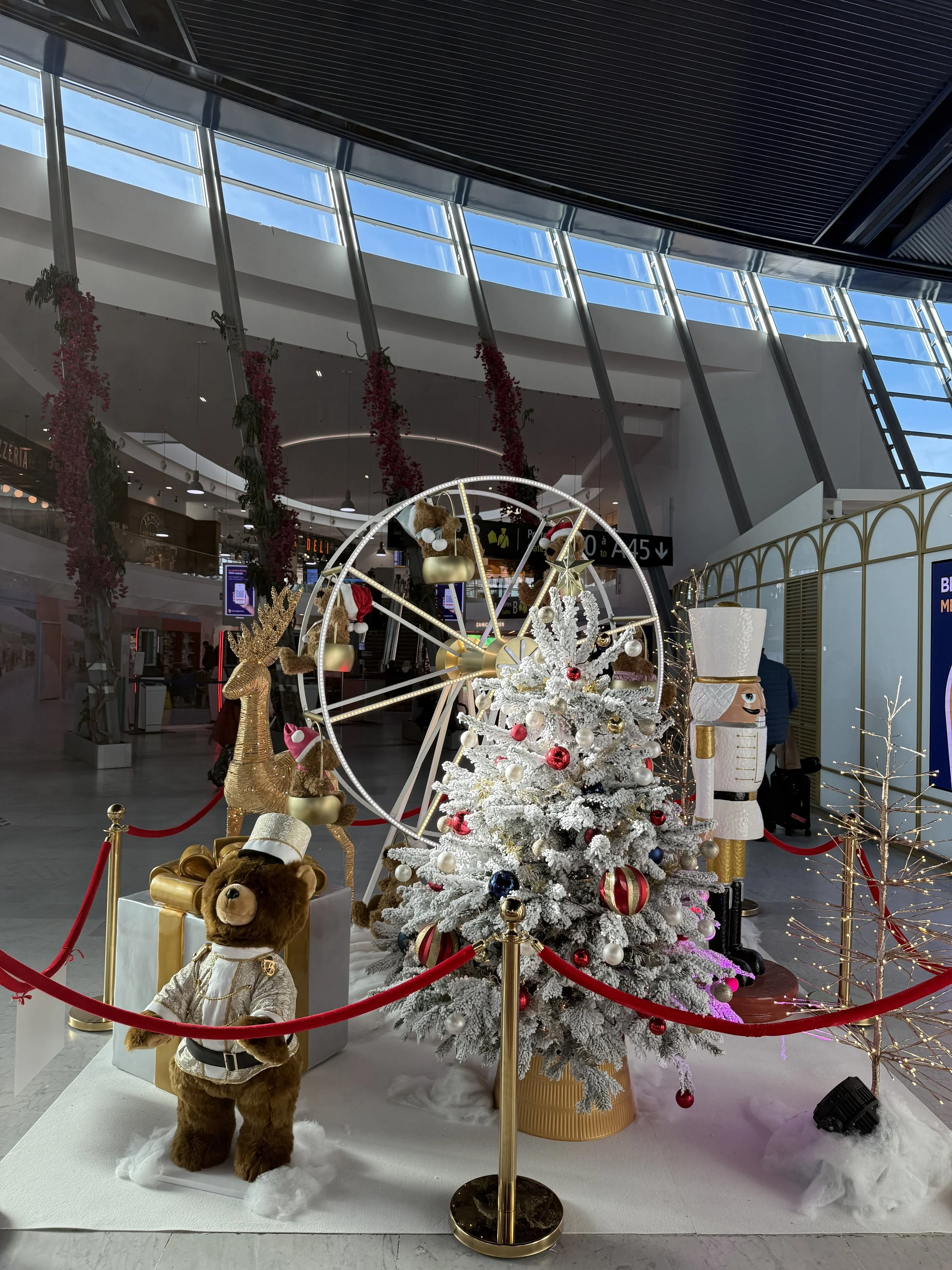 Décor de Noël avec un sapin blanc décoré, des ours en peluche vêtus de costumes de fête, une grande figurine d'ours en peluche en uniforme et des décorations de Noël entourées de cordes rouges dans un aéroport.