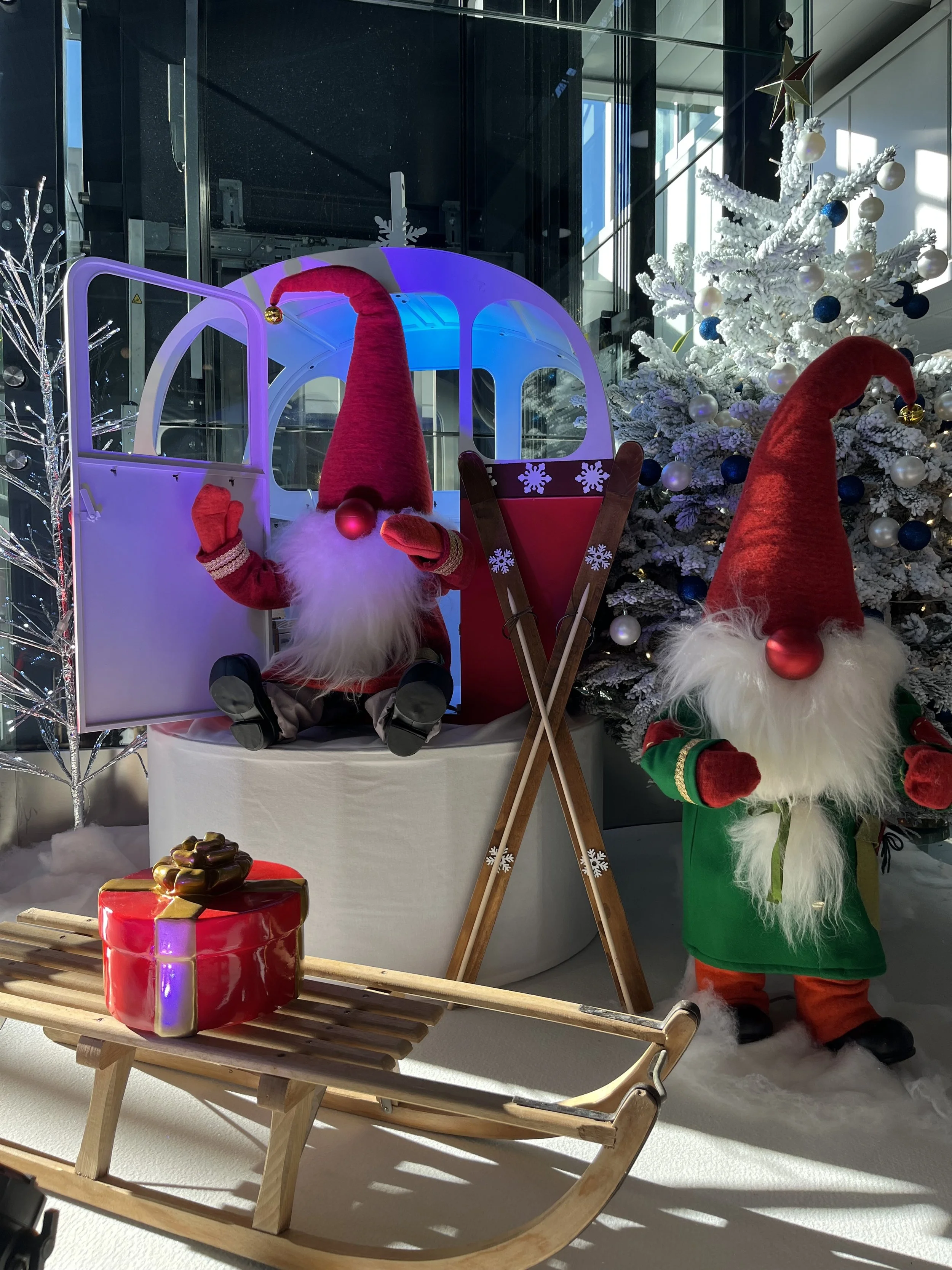 Décor de Noël mettant en scène deux Pères Noël, l'un assis dans un traîneau blanc et l'autre debout, tous deux vêtus de costumes et de bonnets rouges. Un sapin de Noël orné de décorations bleues et blanches se dresse derrière eux. 