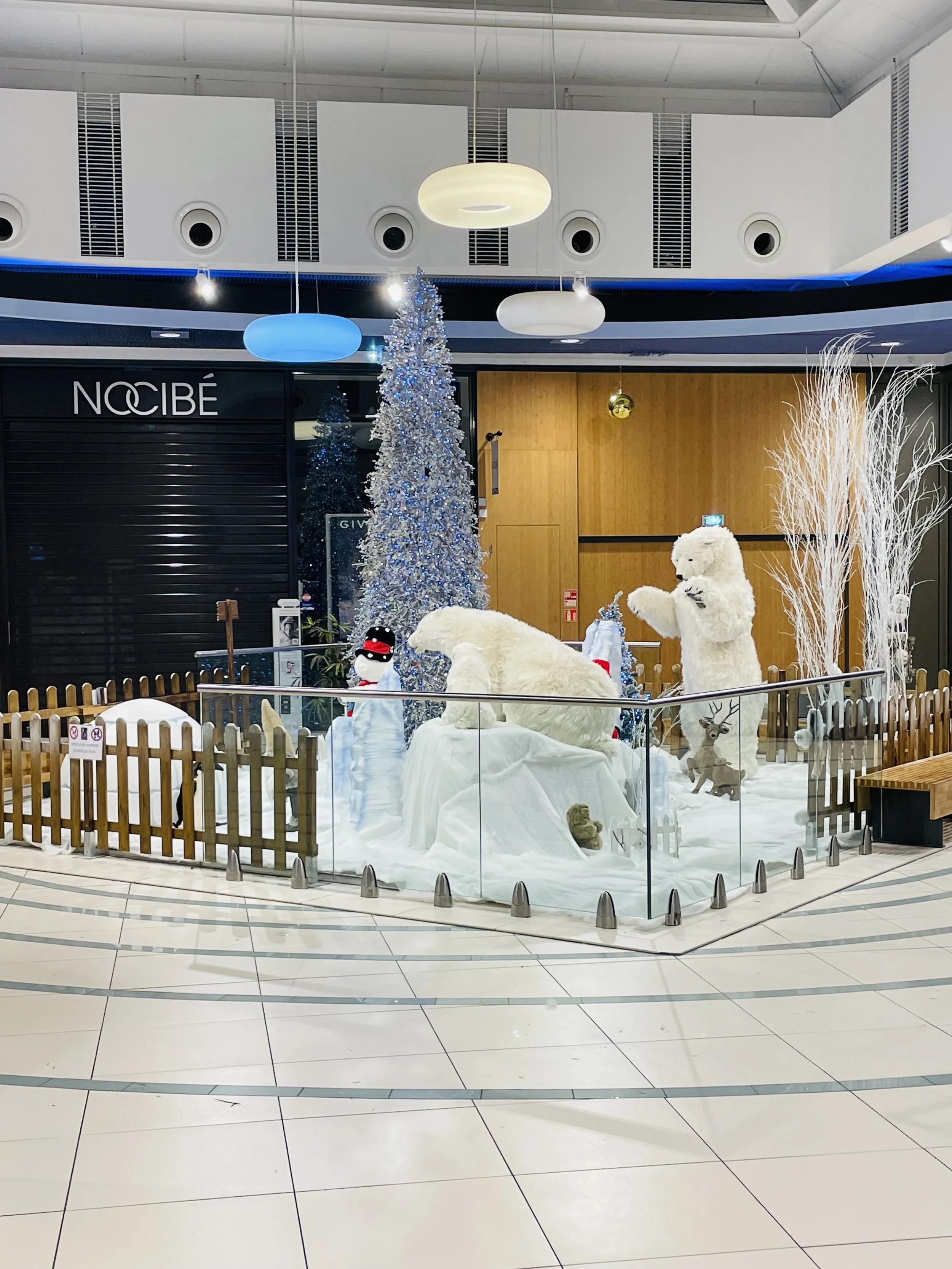 Décoration intérieure de Noël comprenant des sculptures d'ours polaires blancs automates, un sapin décoré, des bonshommes de neige, des rennes et des branches blanches, le tout entouré d'une petite clôture en bois. dans un centre commercial.
