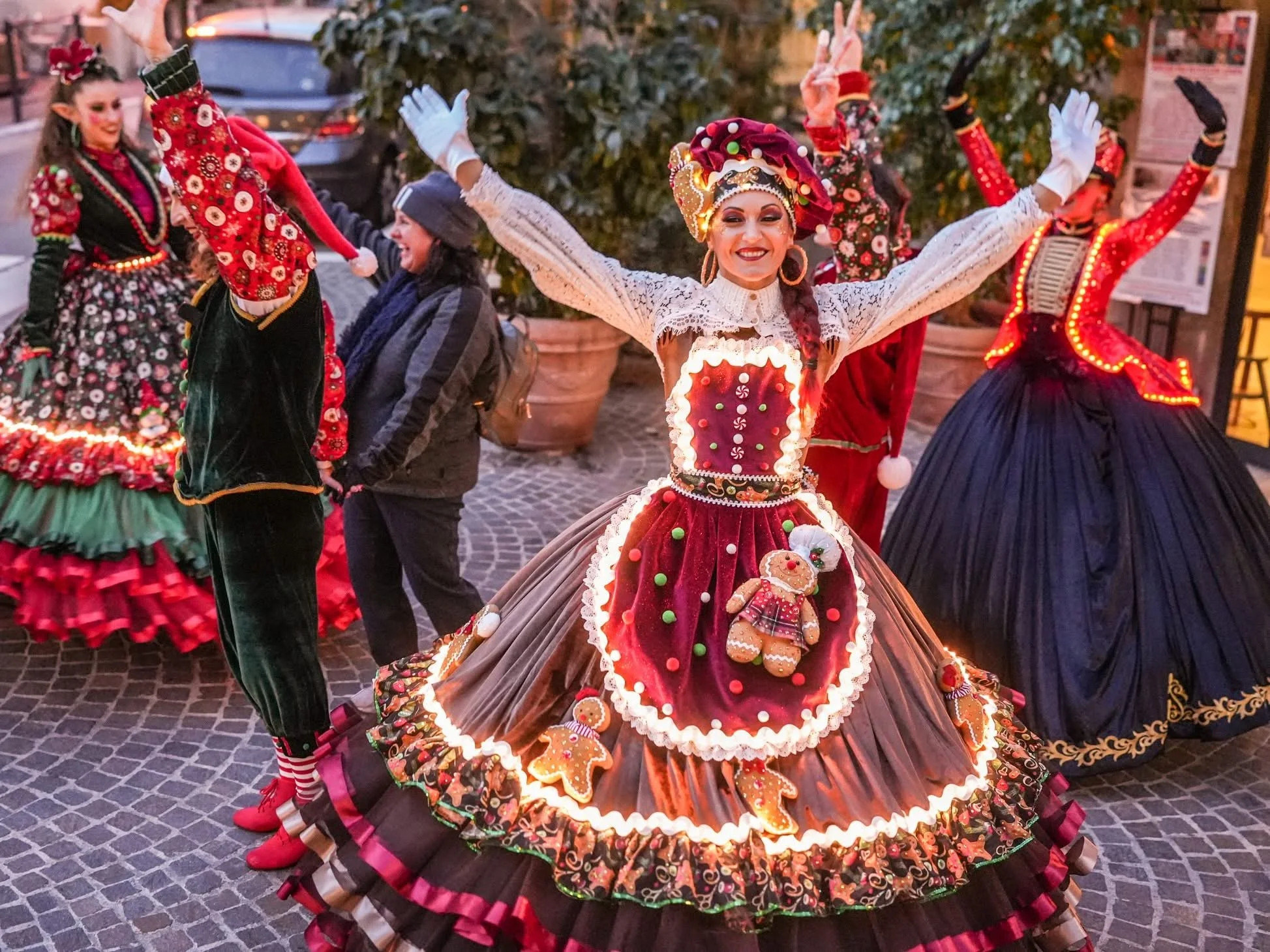 Fête de Noël avec des personnes en costumes de Noël, notamment une femme au centre portant une robe et un chapeau décorés de motifs de pains d'épices, entourée d'autres personnes en costumes festifs, dans une ambiance lumineuse extérieure en soirée. 