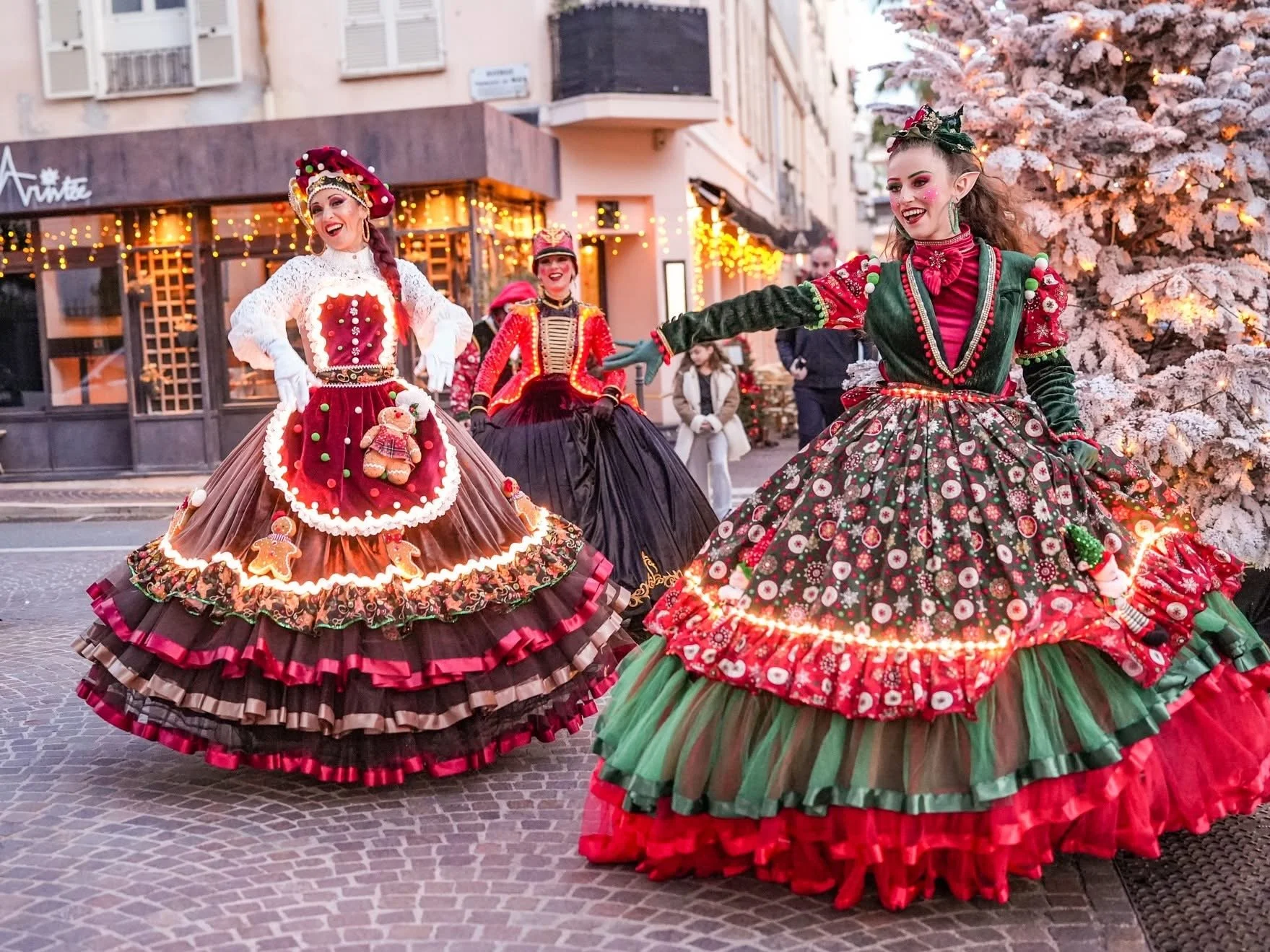 Des personnes déguisées en lutins de Noël dans une rue décorée pour Noël, avec un sapin enneigé en arrière-plan. Dans les alpes maritimes 06 à cannes