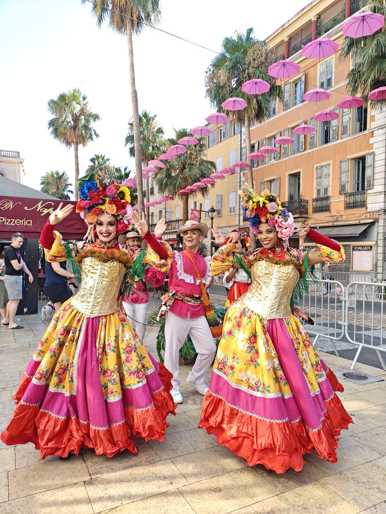 Trois danseurs en costumes colorés avec des chapeaux décorés de fleurs, lors d'une parade en plein air, avec des palmiers et un bâtiment orange en arrière-plan.