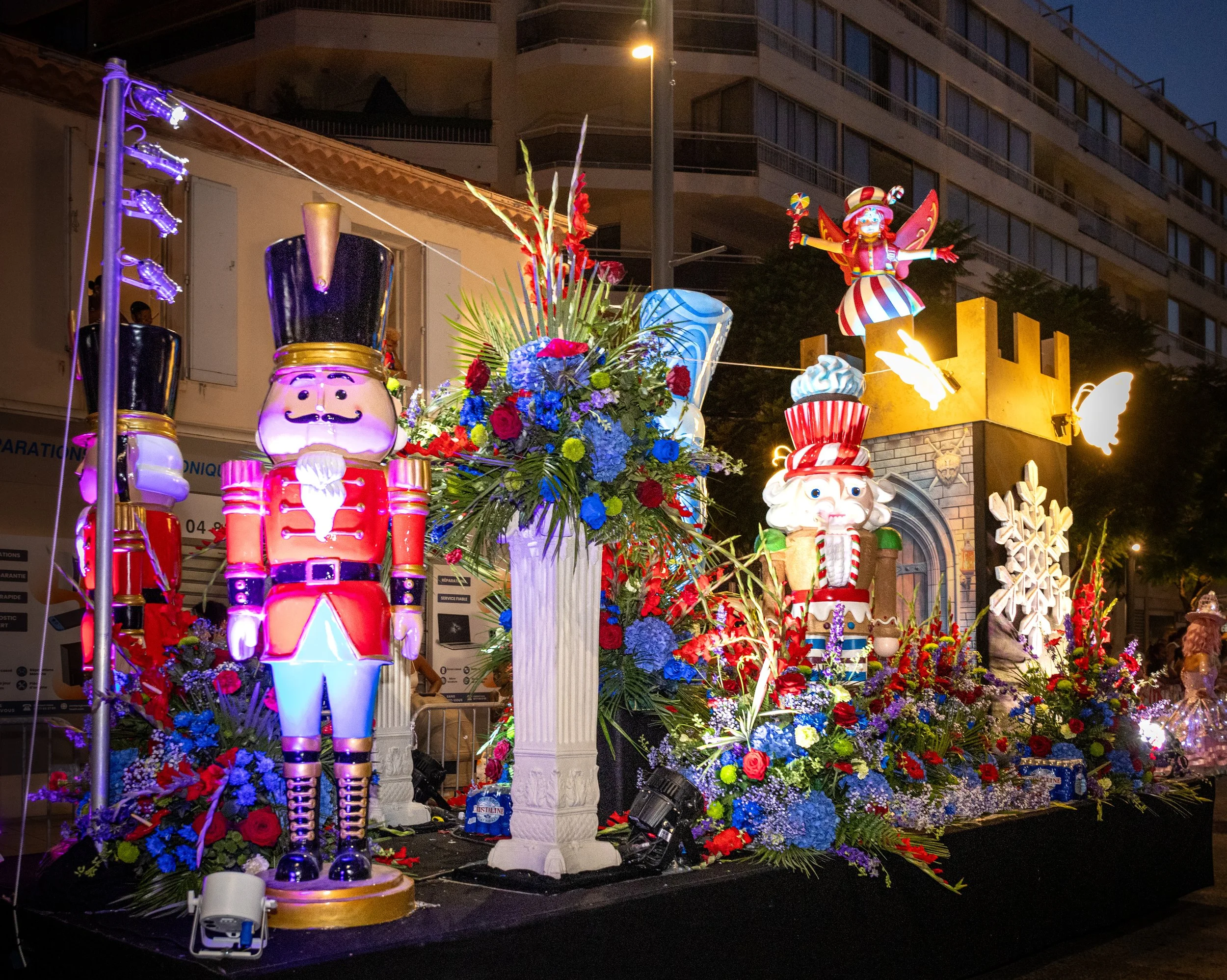 Scène nocturne d'un char festif décoré de grandes figurines de casse-noisettes, de fleurs éclatantes et d'un personnage de clown féerique et fantaisiste, avec des bâtiments et des lampadaires en arrière-plan.