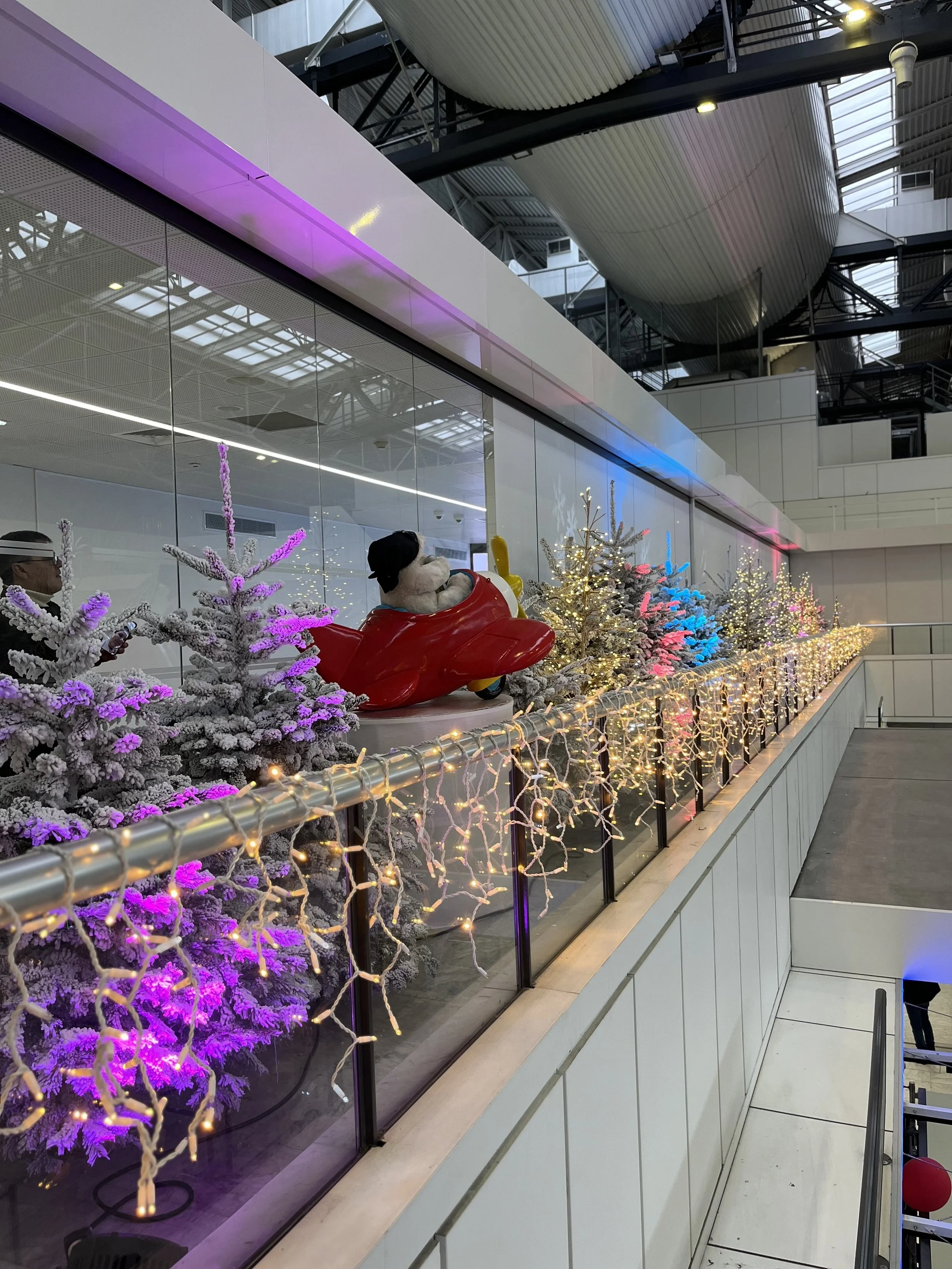 Intérieur d'un aéroport ou d'un terminal avec des sapins de Noël décorés, des illuminations festives et une grande figurine du Père Noël sur un scooter devant une paroi vitrée avec des reflets.