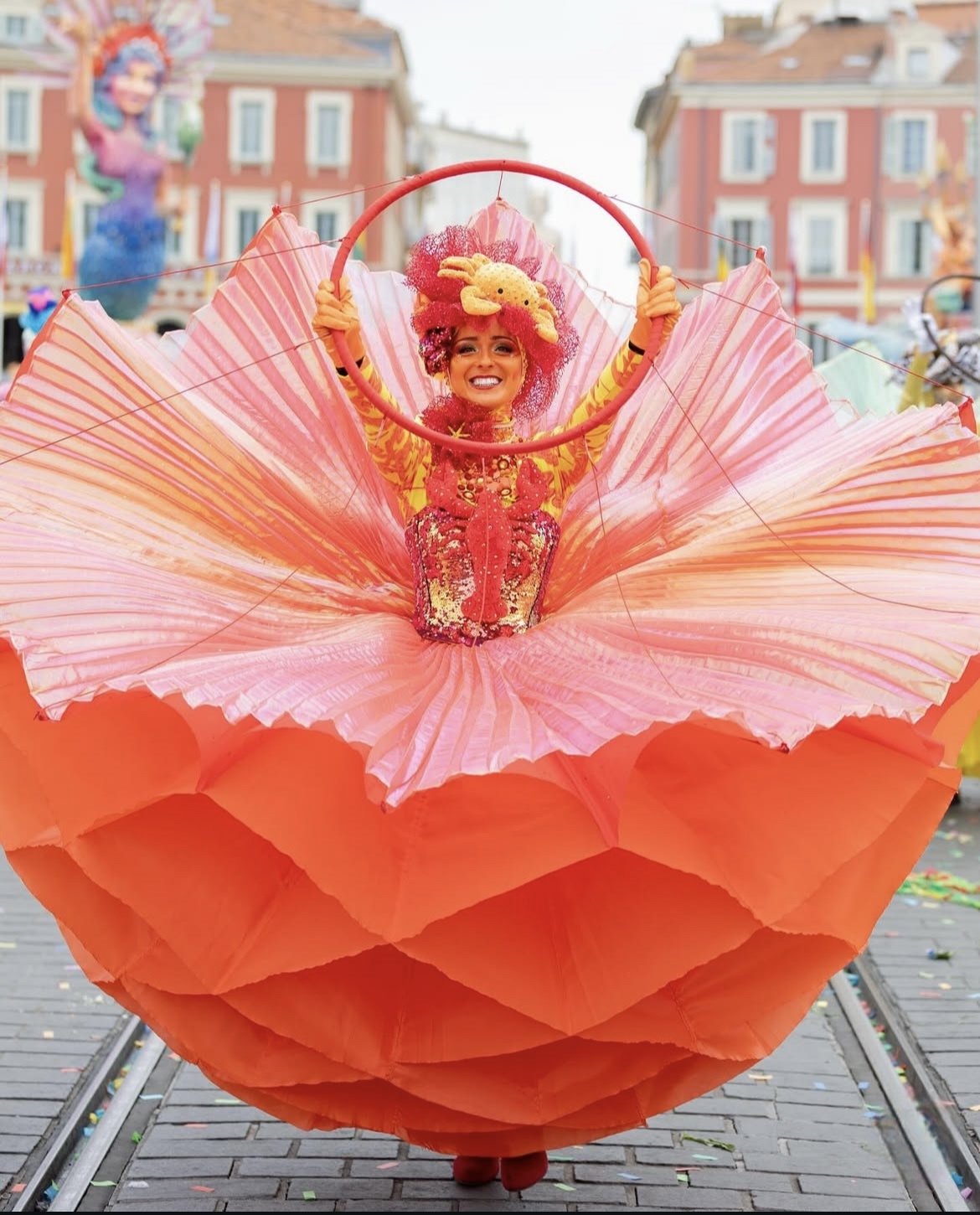 Une femme en costume coloré et festif, avec une tête ornée d’un coq en peluche, se trouve dans une grande structure en papier rose et orange, lors d’un défilé ou carnaval en plein air.