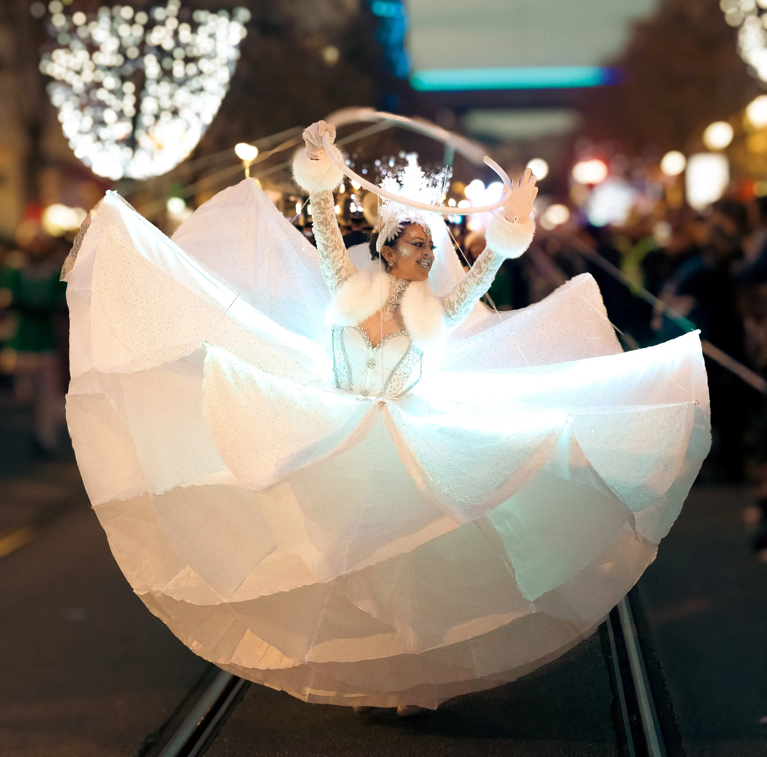 Une femme avec un maquillage brillant et un costume de danseur en blanc, levant les bras lors d'une parade nocturne dans une ville éclairée. Dans les alpes maritimes 06 à nice.
