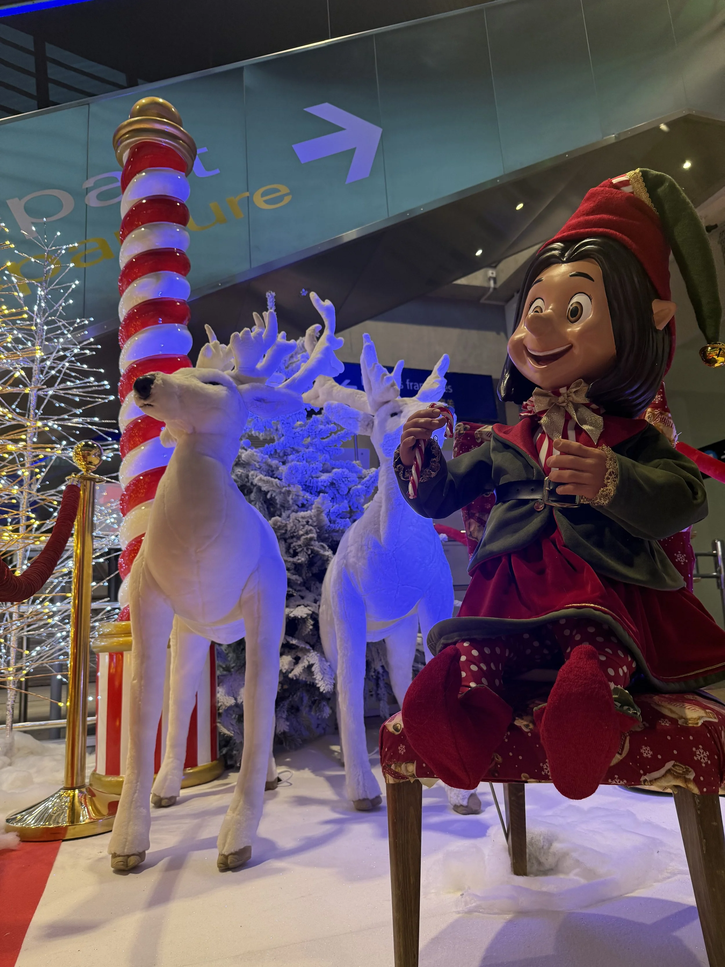 Décor de Noël avec une grande figurine d'elfe, des sculptures de rennes blancs, un sapin de Noël décoré et une canne de Noël rayée, le tout sur un sol enneigé, avec un panneau indiquant la direction.