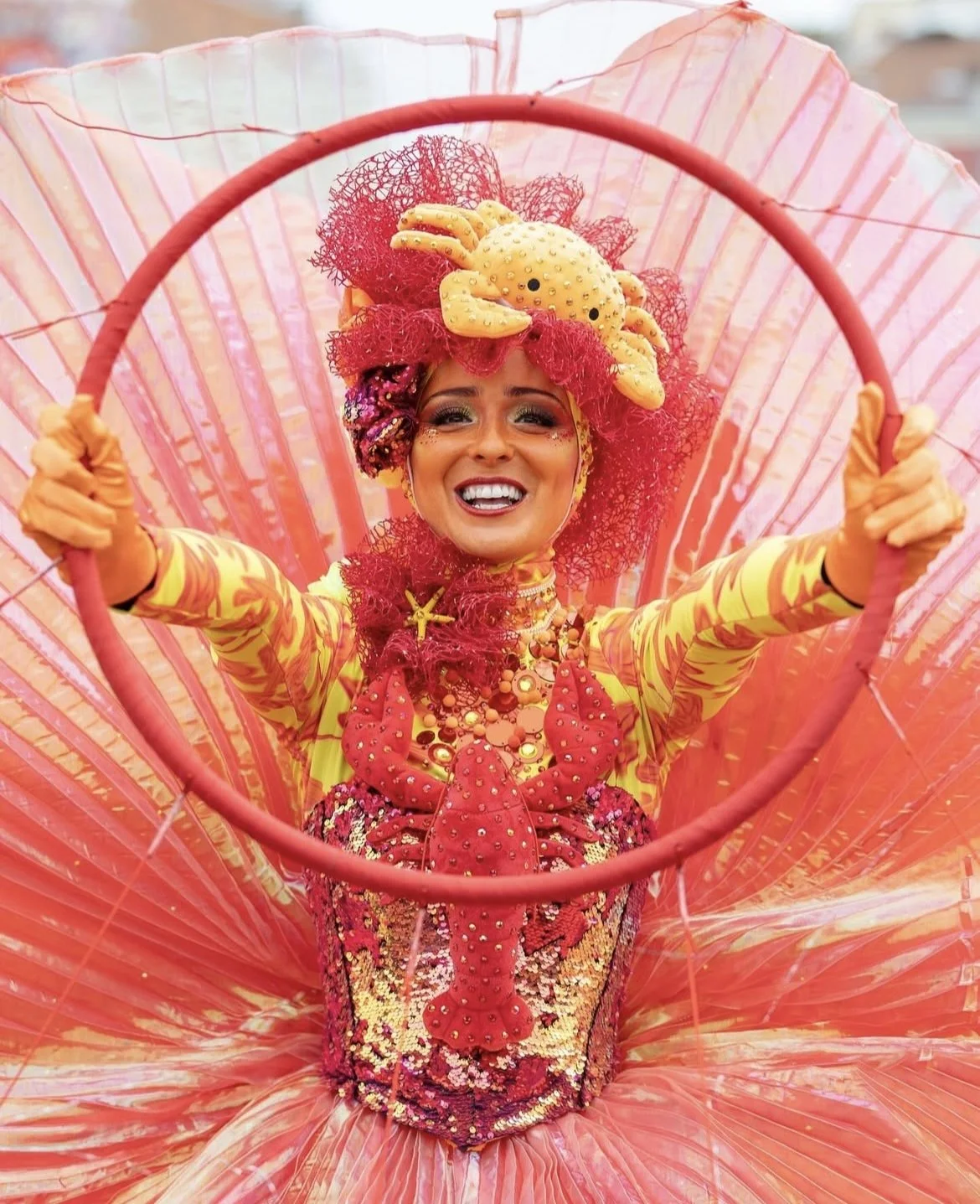 Une femme souriante portant un costume coloré de carnaval avec un thème marin, comprenant un chapeau en forme de crabe avec une créature marine, entourée d'un grand parapluie rose. Elle est vêtue de jaune avec des détails rouges et des accessoires au