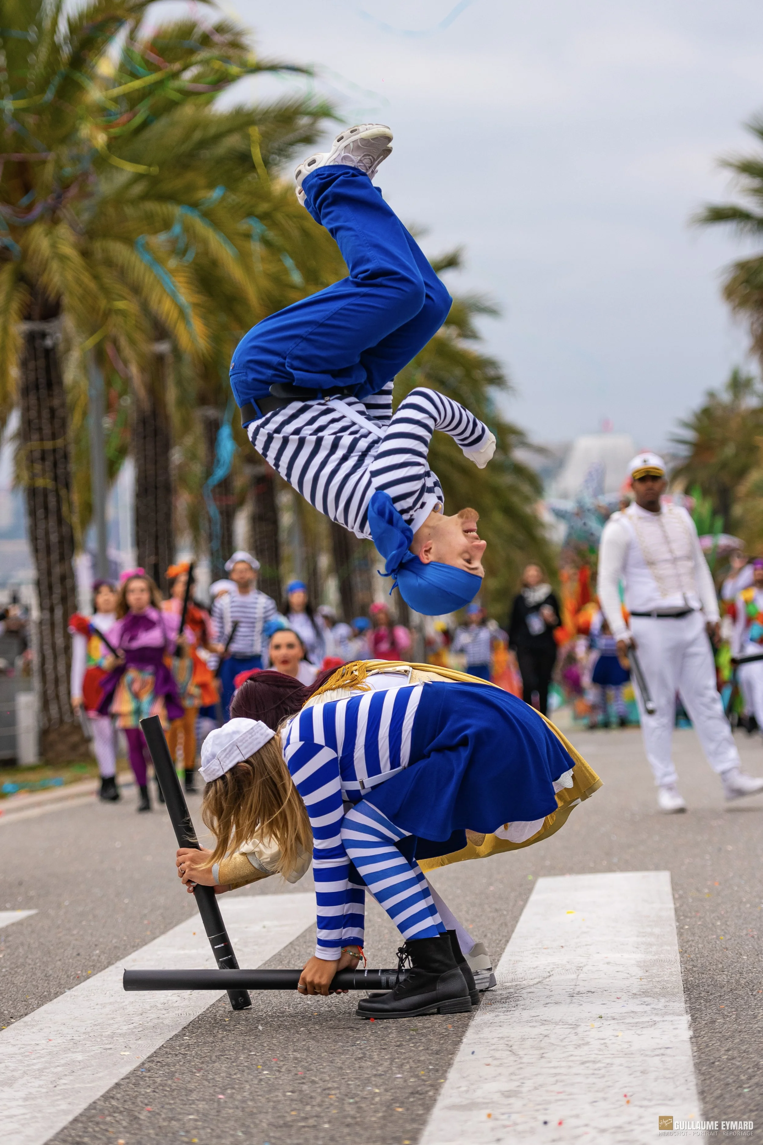 Des enfants déguisés en marins font un spectacle acrobatique lors d'une parade ou d'un festival. Ils sont en équilibre, avec un public de spectateurs en arrière-plan. L'ambiance est festive et colorée.