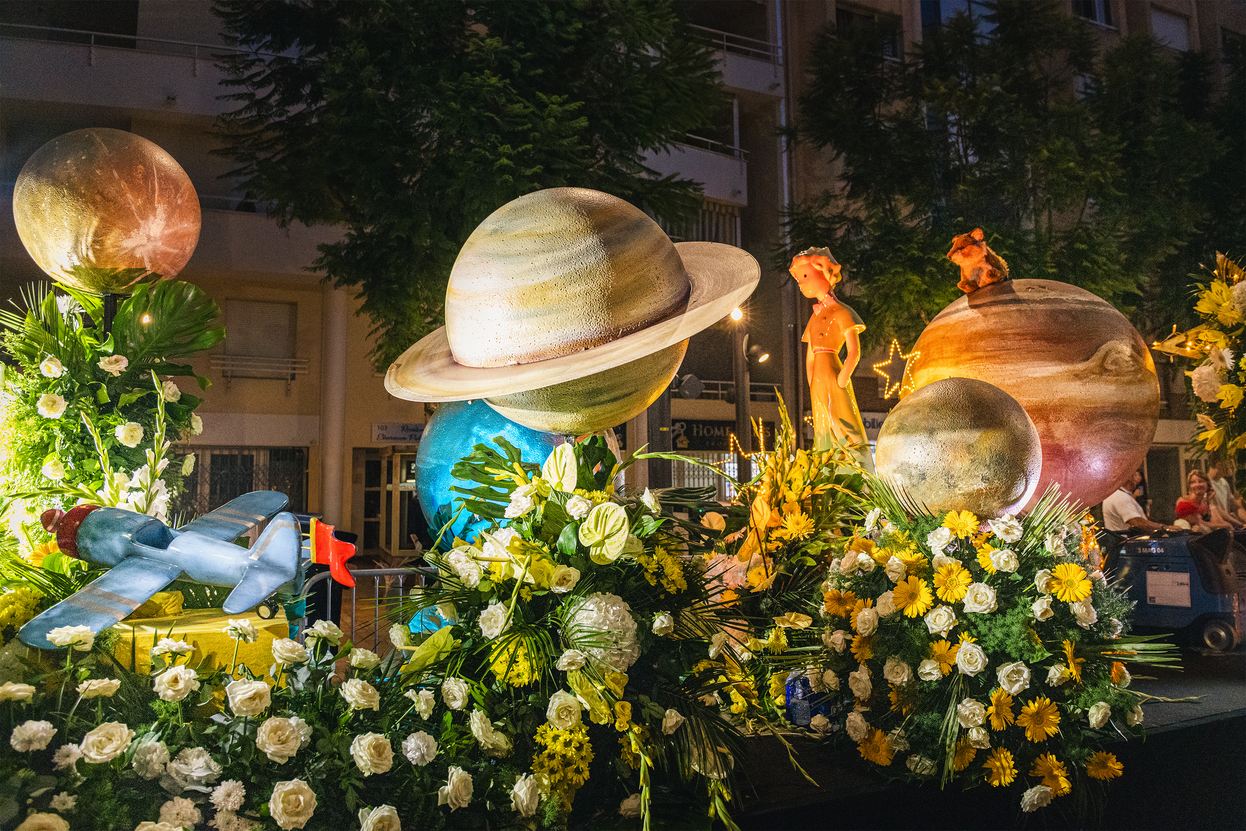Décor d'une parade ou d'un festival avec des figurines et des éléments décoratifs inspirés de l'espace, avec des planètes et des fleurs colorées, éclairés la nuit.