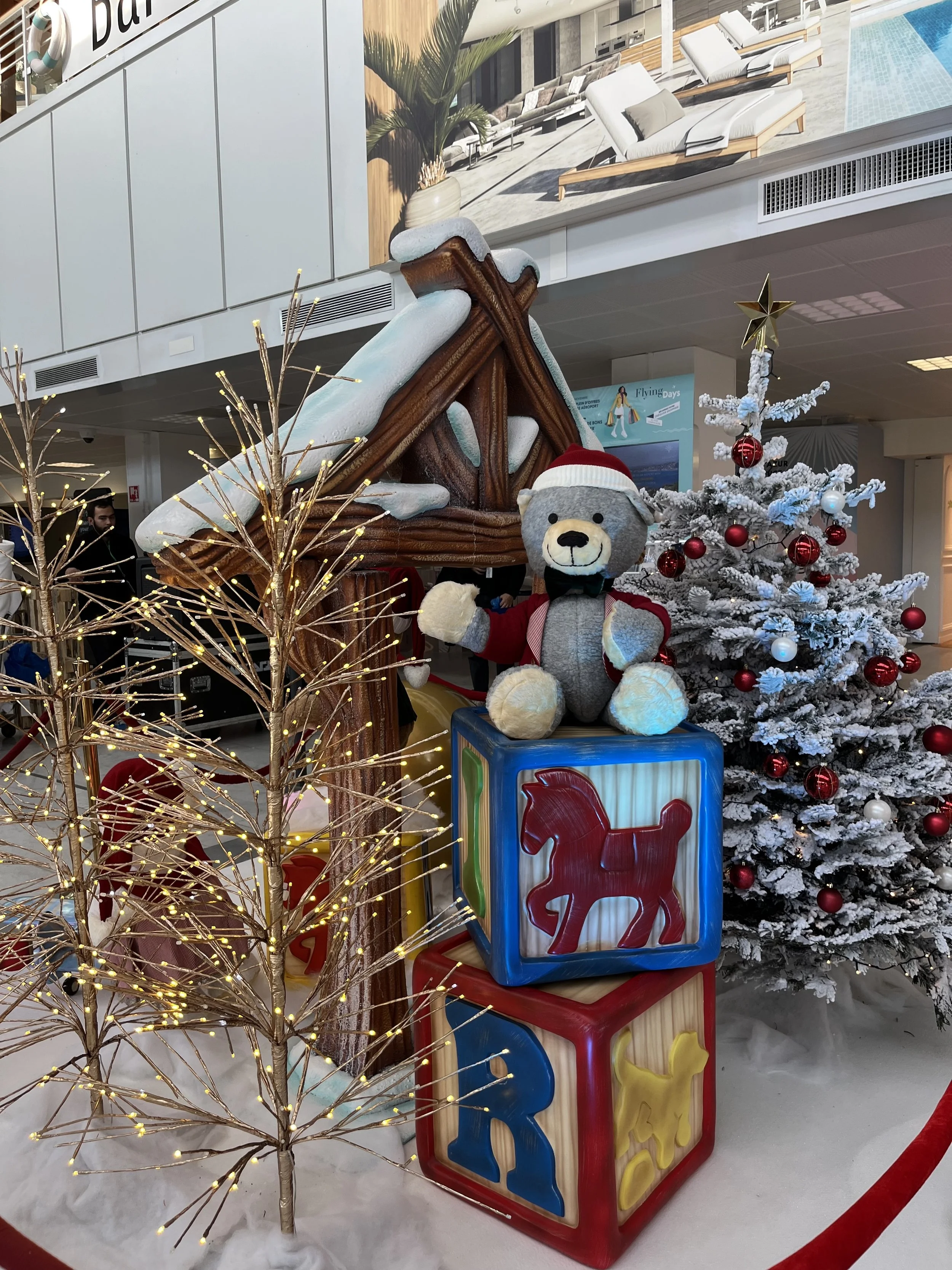 Décor de Noël mettant en scène un sapin artificiel enneigé et décoré de boules rouges et blanches, un ours en peluche portant un bonnet et une veste de Père Noël, ainsi qu’une maison en pain d’épices en bois recouverte de neige sur le toit. L’ensembl