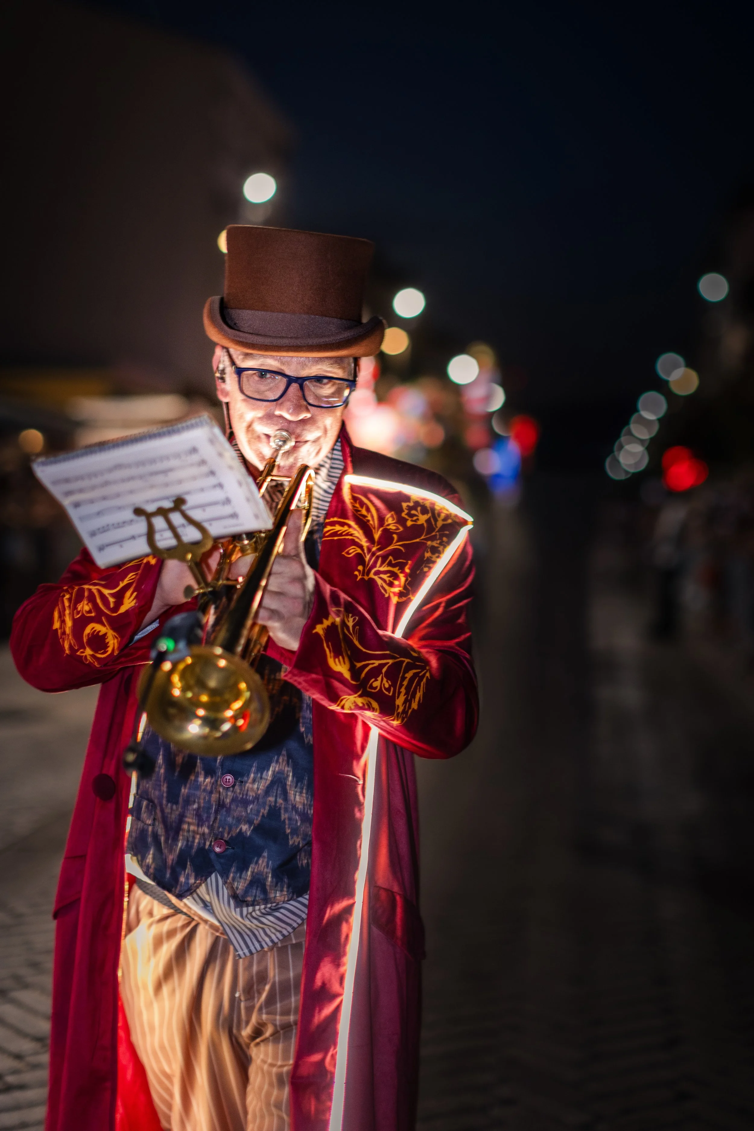 Un homme jouant de la trompette en plein air la nuit, portant des lunettes, un haut-de-forme marron, un blazer en velours coloré à broderies dorées, une chemise à motifs et un pantalon rayé, avec les lumières de la ville et un ciel sombre en arrière-