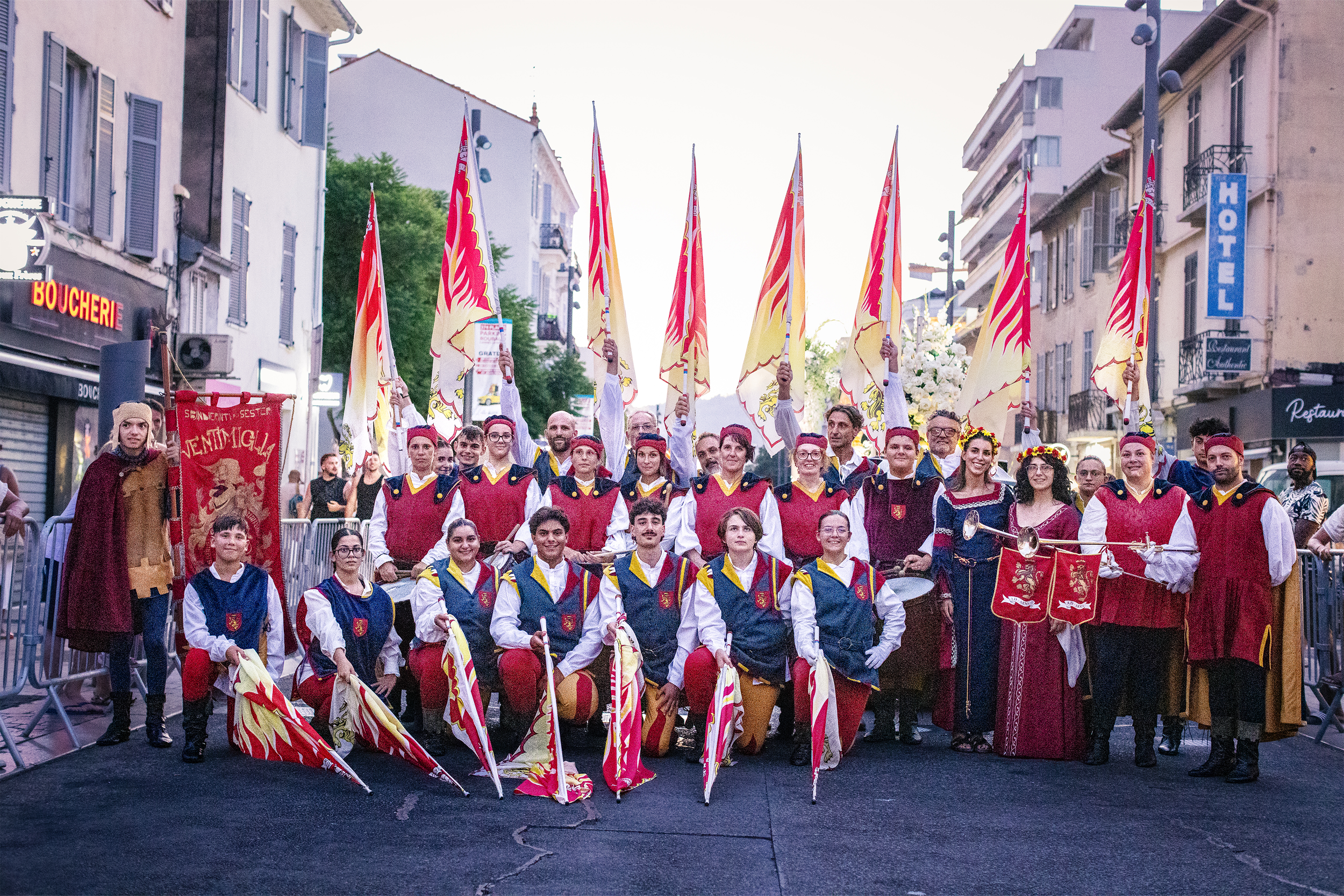 Groupe de personnes en costumes traditionnels avec drapeaux lors d'une parade en ville, sous un ciel clair, avec des bâtiments en arrière-plan.