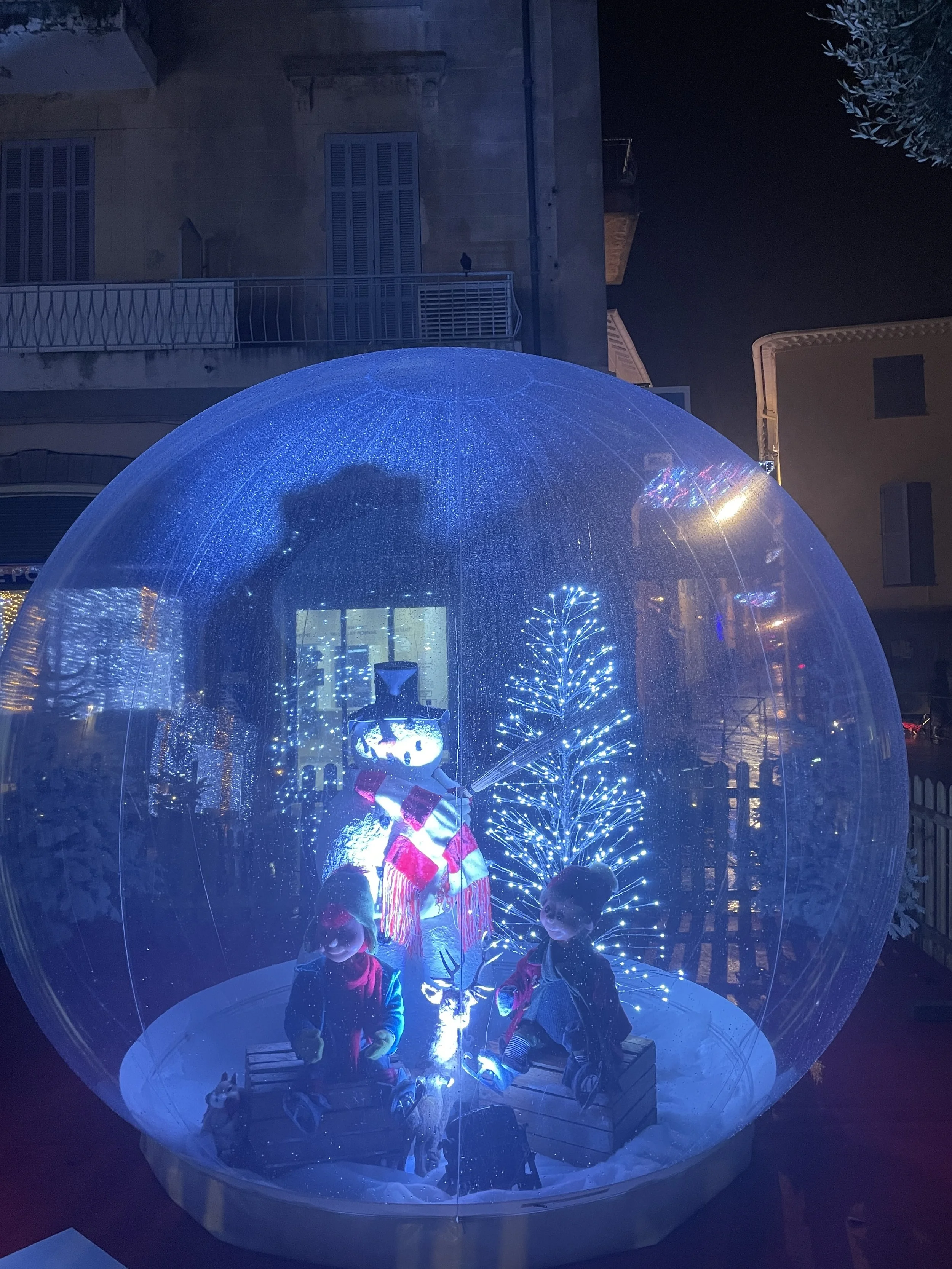 Des enfants à l'intérieur d'un dôme gonflable transparent décoré de lumières bleues, évoquant une décoration de Noël, avec un petit sapin et d'autres décorations de fêtes visibles à l'intérieur.