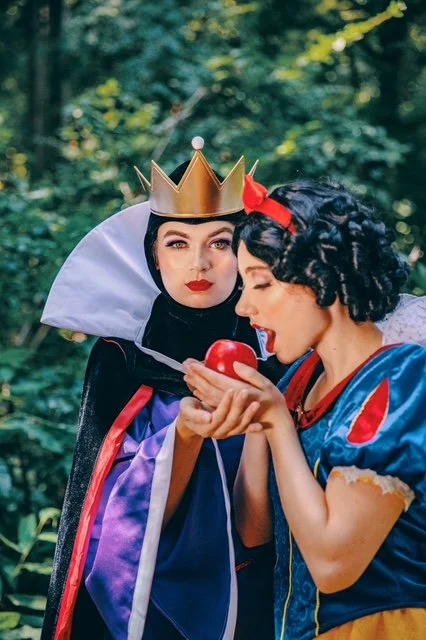 Deux femmes en costumes de contes de fées dans une forêt, l'une portant une couronne et une cape noire, l'autre en robe bleue avec un ruban rouge, tenant une pomme rouge.