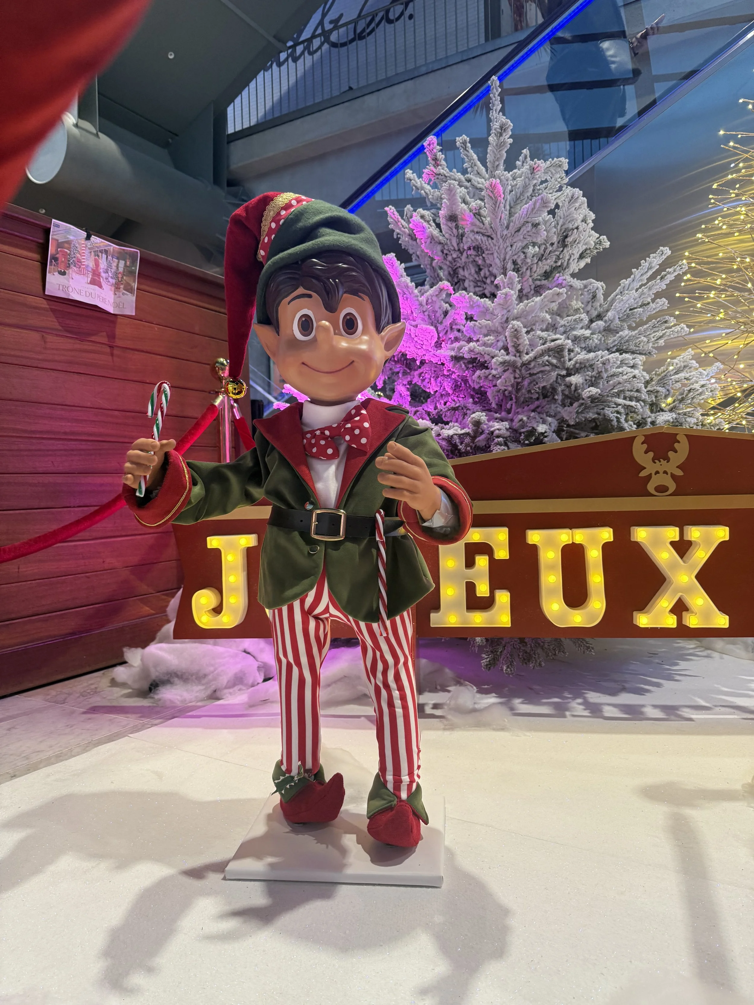 Une décoration de Noël festive mettant en scène un joyeux elfe tenant une canne de Noël, vêtu d'une tenue verte et rouge avec un pantalon rayé et des pantoufles rouges, se tenant devant un décor enneigé avec un sapin de Noël enneigé illuminé