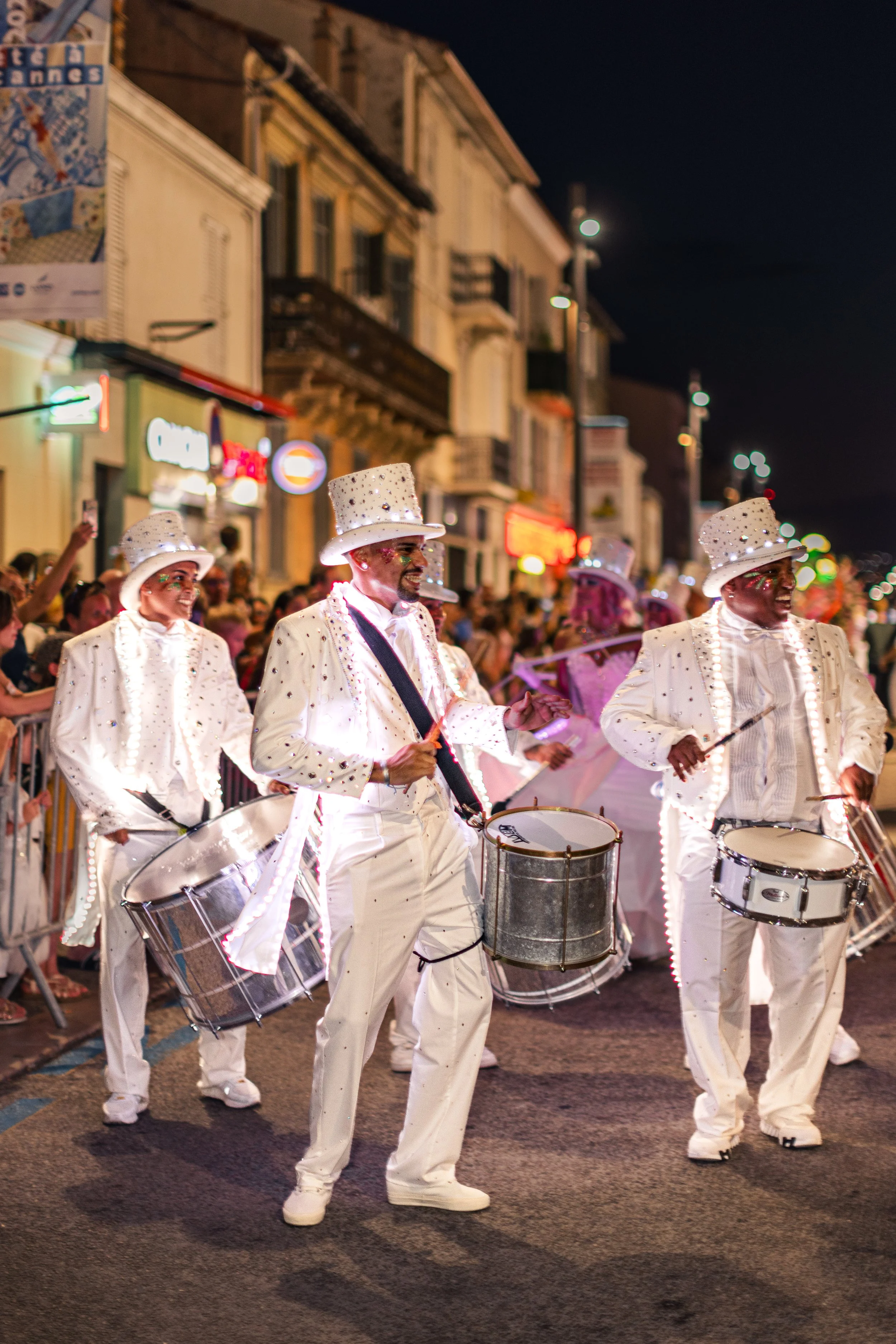 Un groupe de musiciens vêtus de costumes blancs à bordures lumineuses et de chapeaux assortis, jouant du tambour et des percussions lors d'un défilé nocturne dans une rue de la ville, sous le regard d'une foule.