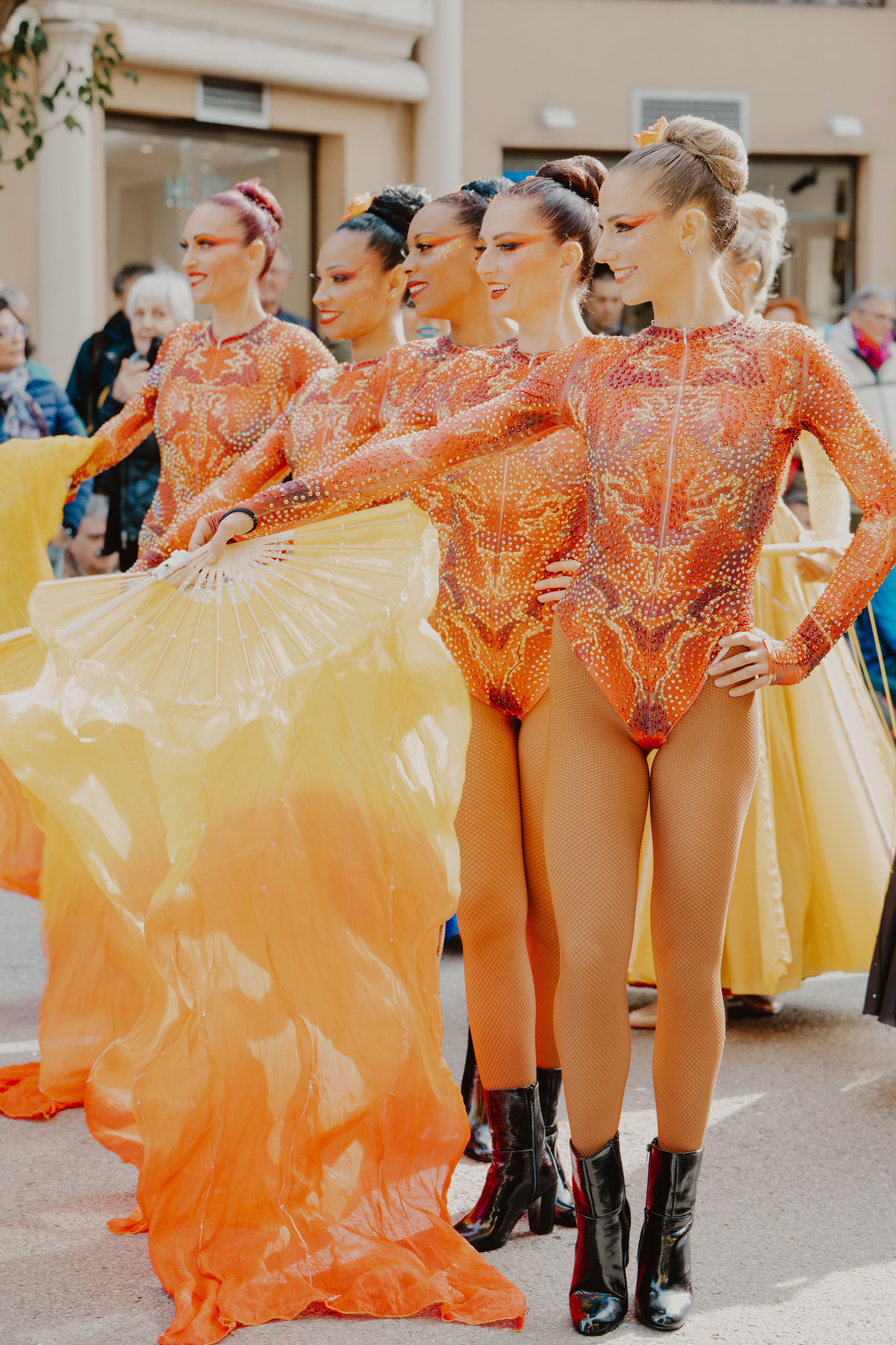 Groupe de danseuses en costumes orange et noir avec des jupes en tulle orange, en train d'effectuer une performance dans la rue, avec un public en arrière-plan.