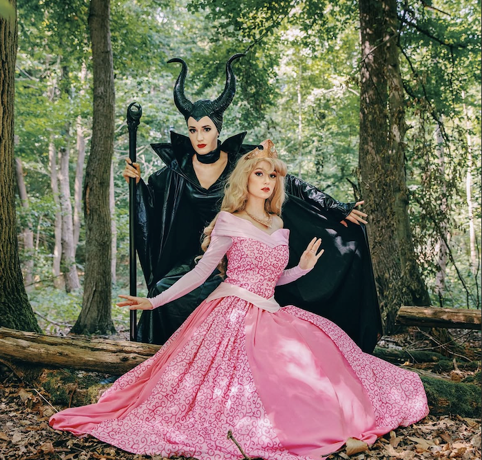 Deux femmes déguisées en personnages de conte de fées dans une forêt, l'une en sorcière Maléfique avec des cornes noires et robe noire, l'autre en princesse en robe rose, portant une couronne.