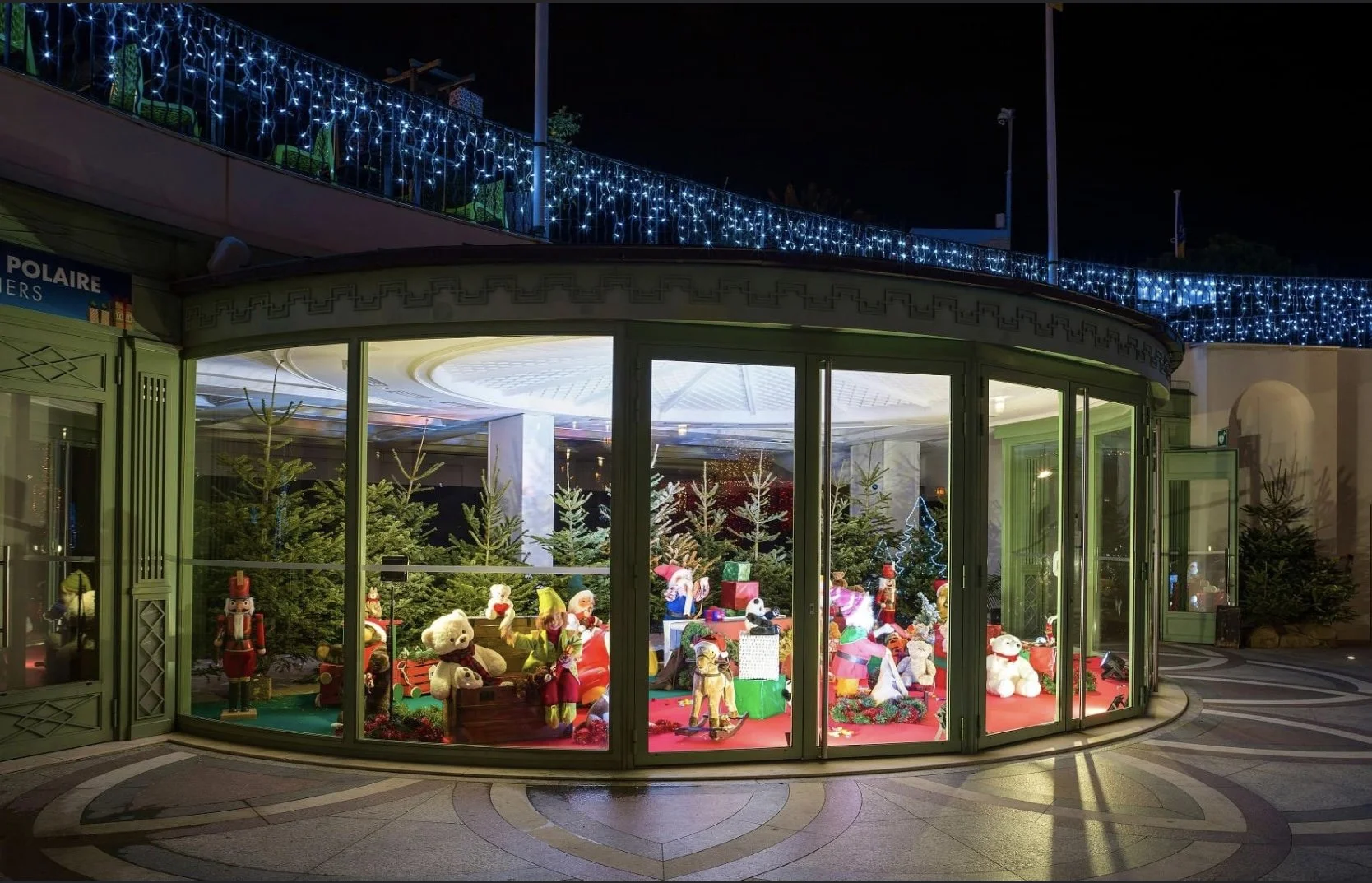 Décorations de Noël sous une structure vitrée, avec des peluches, des ours en peluche et des décorations de Noël, agrémentées de sapins et de cadeaux, illuminées par un éclairage festif la nuit.