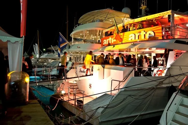 Scène nocturne dans un port de plaisance avec de grands yachts et bateaux, des gens qui discutent sur les ponts, illuminés de lumières colorées, et une enseigne orange bien visible portant le mot « arte » sur l'un des yachts.