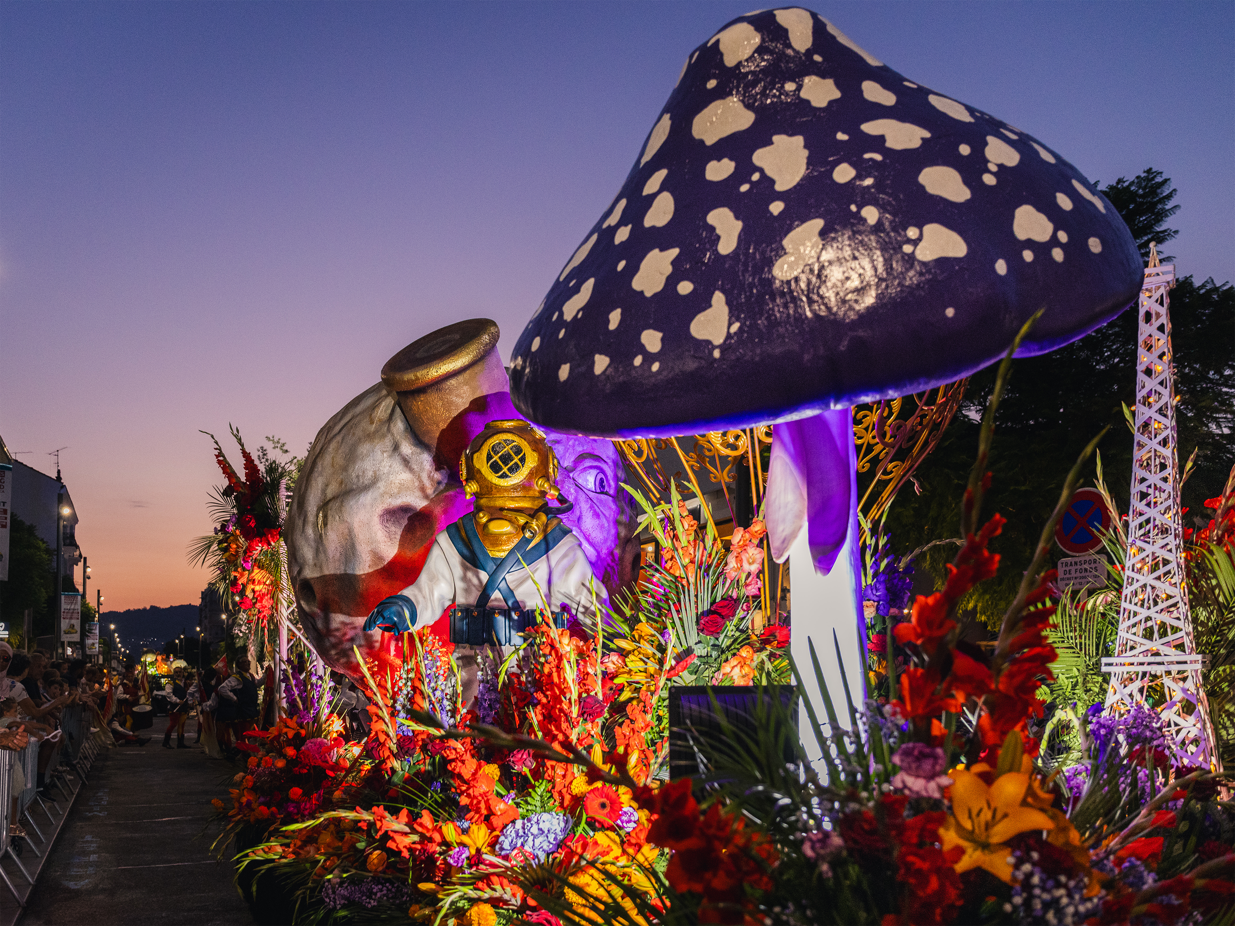 Une grande sculpture représentant un champignon de couleur noire avec des taches blanches, entouré de fleurs colorées lors d'un festival ou parade en plein air en soirée.